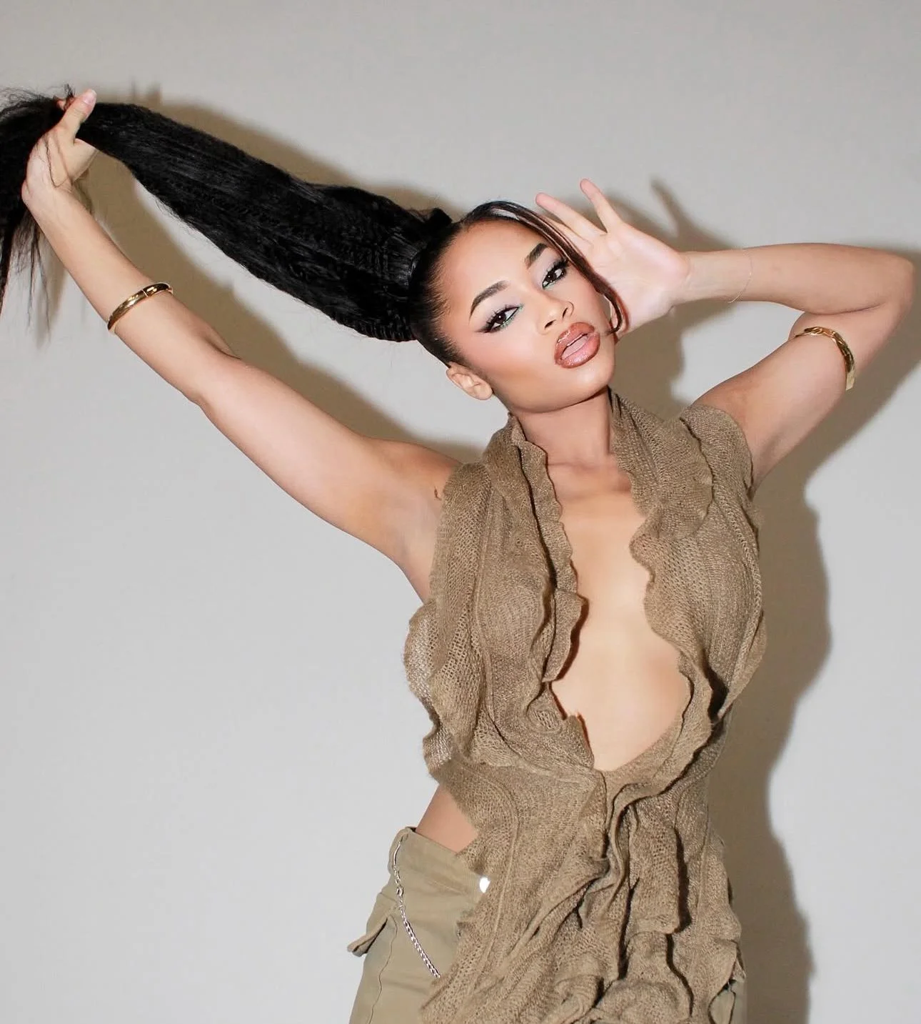 A woman with dark braided hair is posing with one arm raised, holding her hair, and the other hand near her head. She has makeup with winged eyeliner and glossy lips, and is wearing a ruffled, open-front top and khaki pants with a chain accessory.