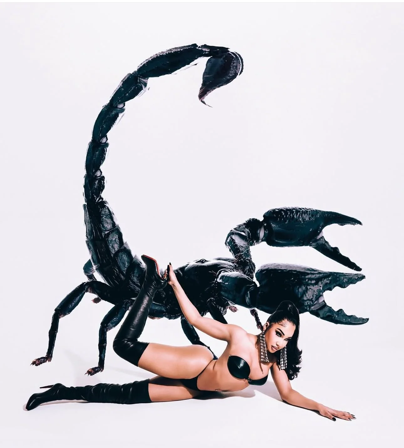 A woman lying on the floor in a provocative pose, wearing black thigh-high boots, black leather shorts, and jewelry, with a giant black sculpture of a scorpion behind her.