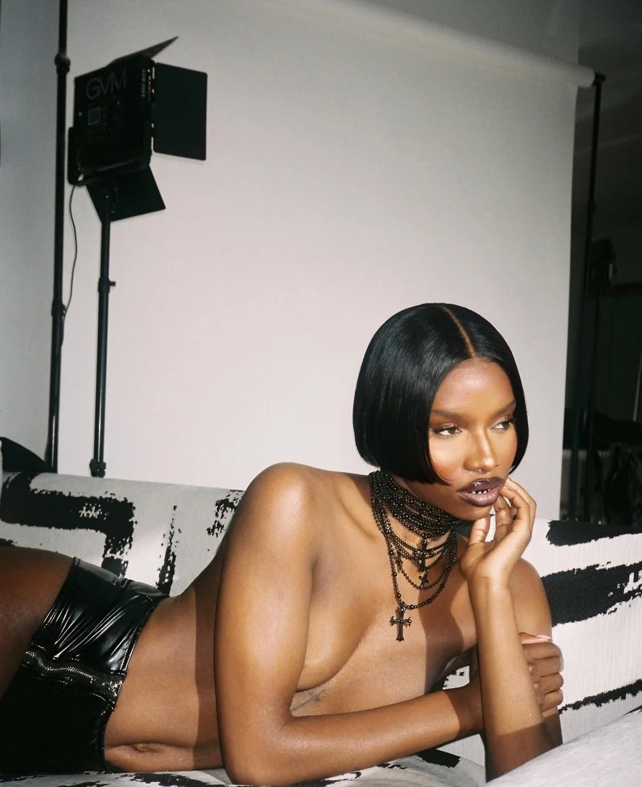 A woman with straight black hair, dark lipstick, and multiple layered necklaces with crosses, lying on a black and white patterned couch in a photography studio with professional lighting equipment.