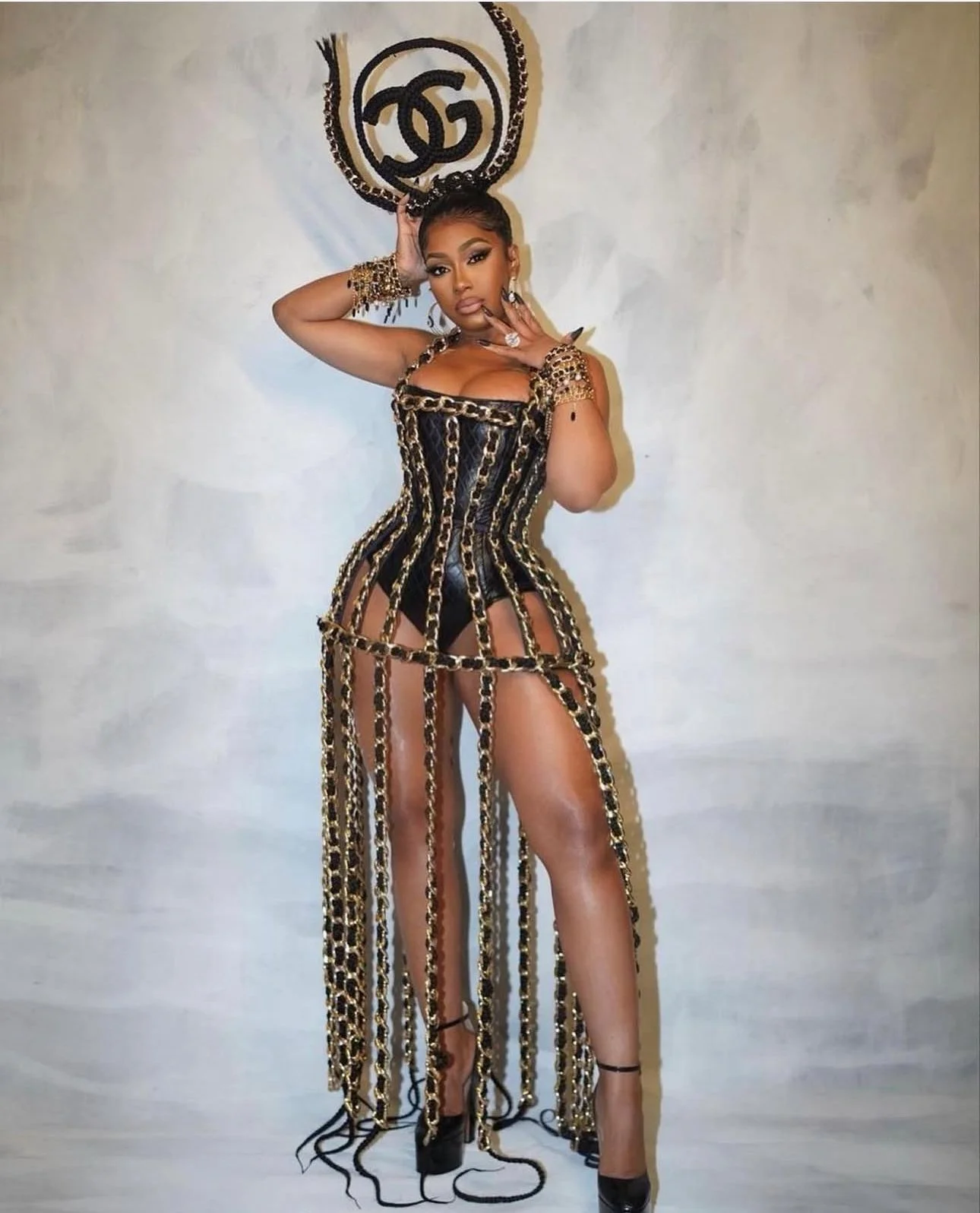 Woman wearing a black and gold chain dress with high heels, standing in front of a wall with a chanel logo on it.