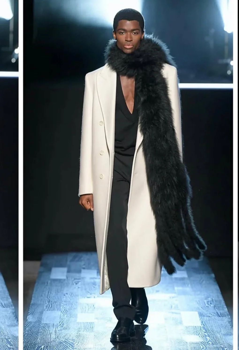 Model walking on runway in black outfit, beige coat, and black fur scarf, with dark background and floor lighting.