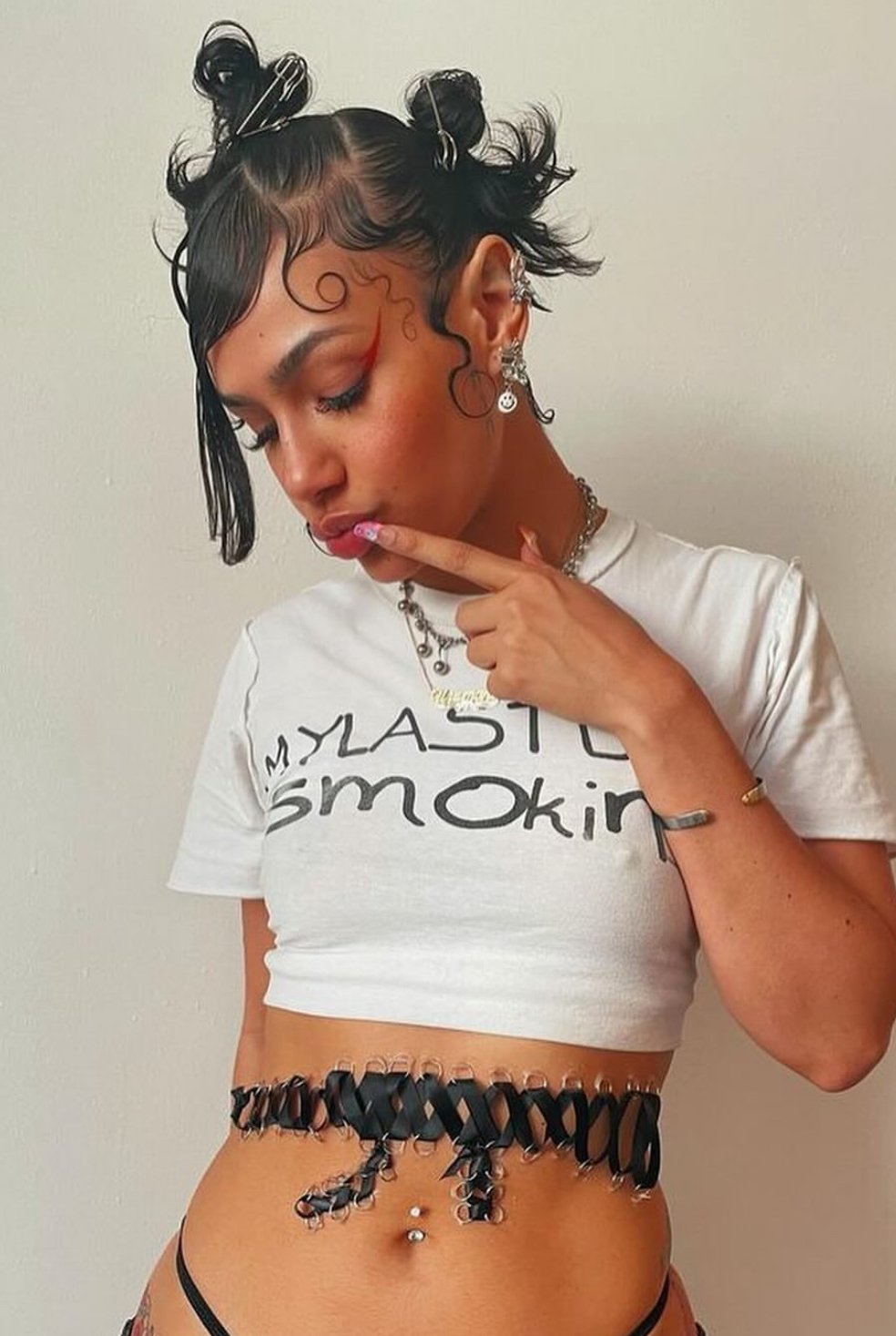 A young woman with short, curly hair styled with hairpins and decorative curls, wearing multiple earrings, a necklace, and a white crop top with the text 'MY LAST smokiN'. She has colorful makeup and is touching her lips with her finger, posing against a plain background.