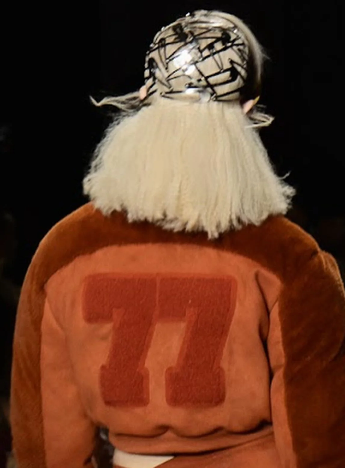 Back of a person wearing a brown sports jacket with a large red '77' on the back and a white beanie with black lines.