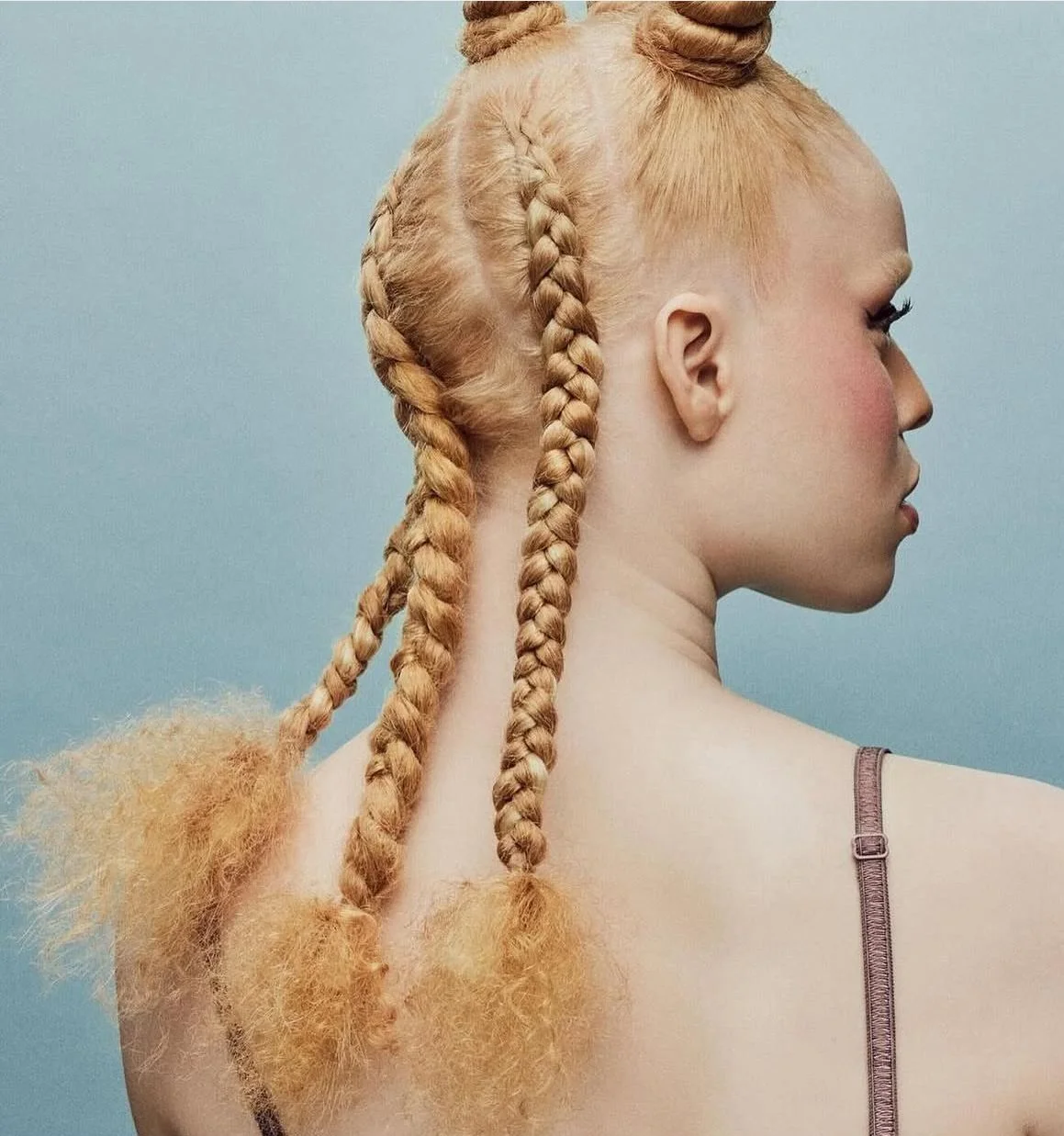 Side profile of a woman with blonde hair styled in multiple braids and fluffy sections, set against a blue background.