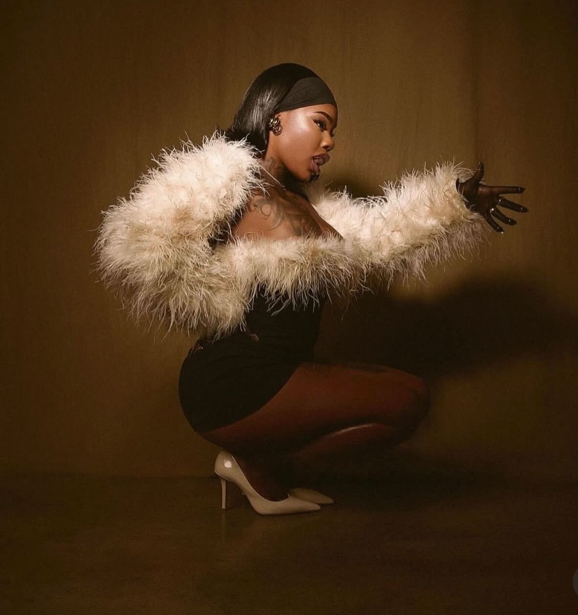 A woman with dark hair in a ponytail wearing a black headband, a large cream-colored feathered coat, black dress, beige high heels, squatting against a plain brown background, with one arm extended outward.