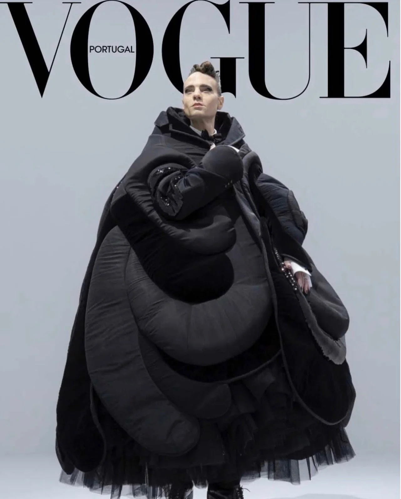 A person dressed in dark, avant-garde fashion with inflated, voluminous clothing, posed against a light gray background at a Vogue Portugal photoshoot.