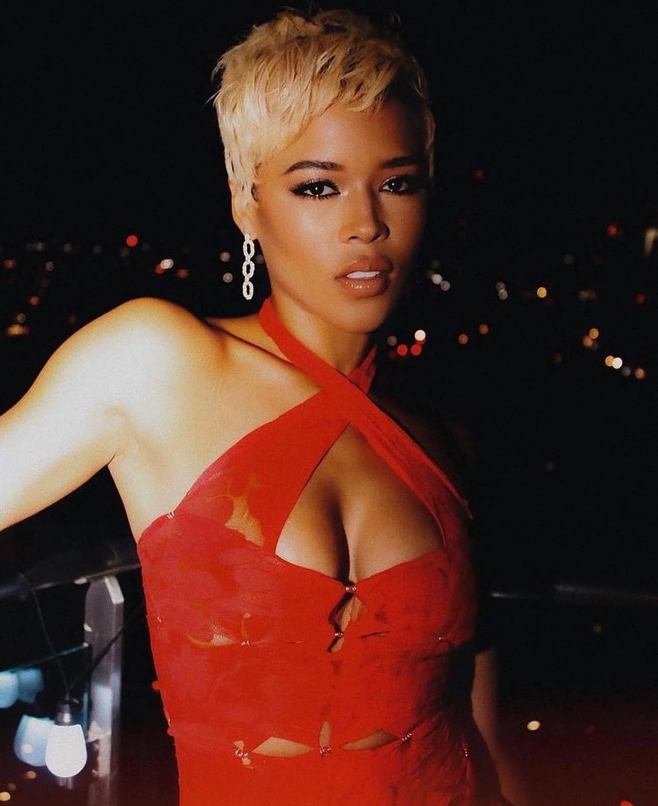 A woman with short blonde hair wearing a revealing red dress and dangling earrings, posing at night with city lights in the background.