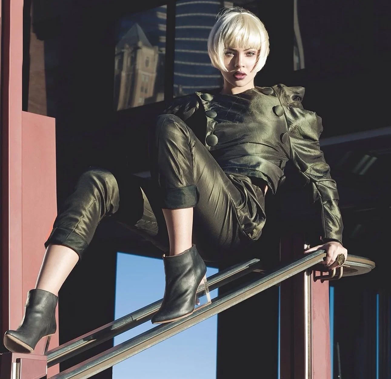 A woman with short blonde hair in a shiny military green outfit and high-heeled ankle boots, sitting on a metal railing outside a building with glass windows.
