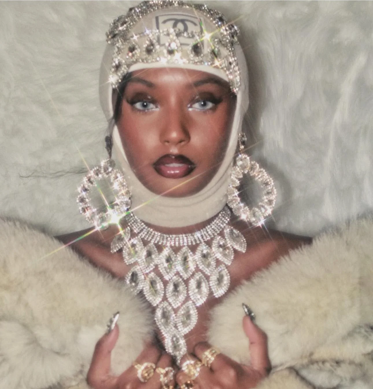 A woman with dark lipstick and eye makeup wearing a white head covering with a large Chanel logo and sparkling rhinestones, surrounded by jewelry including a diamond necklace, earrings, and rings, against a light background.