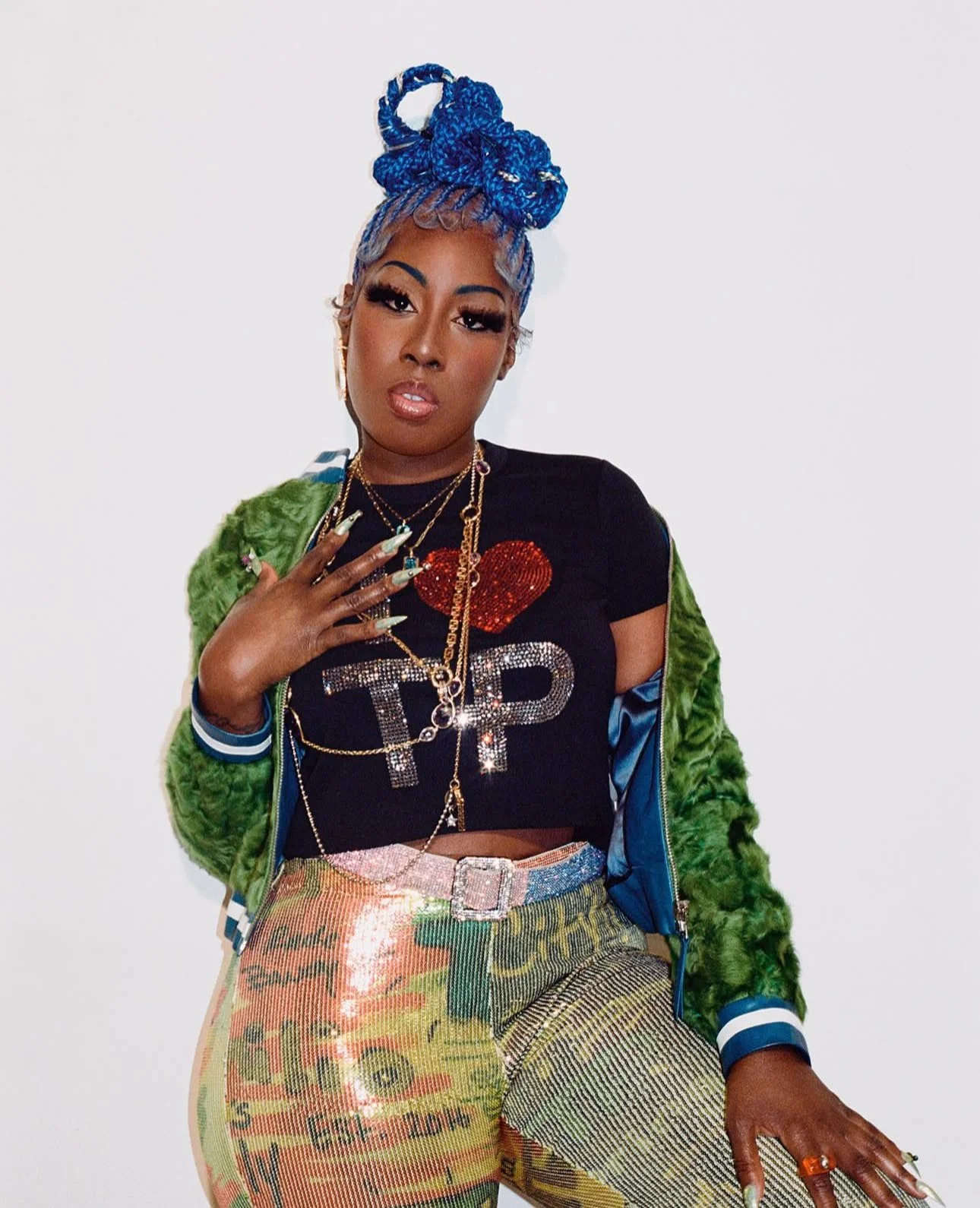 A woman with blue braided hair styled in a bun, wearing a black crop top with a red heart and 'I ❤️ T P' text, layered with jewelry including necklaces, rings, and earrings, and a green fuzzy jacket draped over her shoulders. She is also wearing colorful fabric pants with printed graphics and phrases, standing against a plain white background.