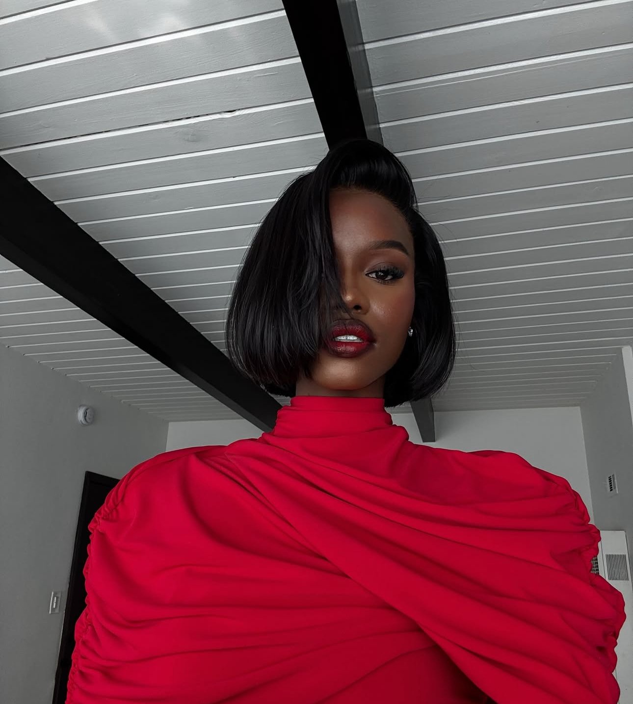 A woman with dark hair styled in a bob, wearing dark makeup and red lipstick, dressed in a red, ruched high-neck top, standing indoors with a white slatted ceiling and a black beam overhead.
