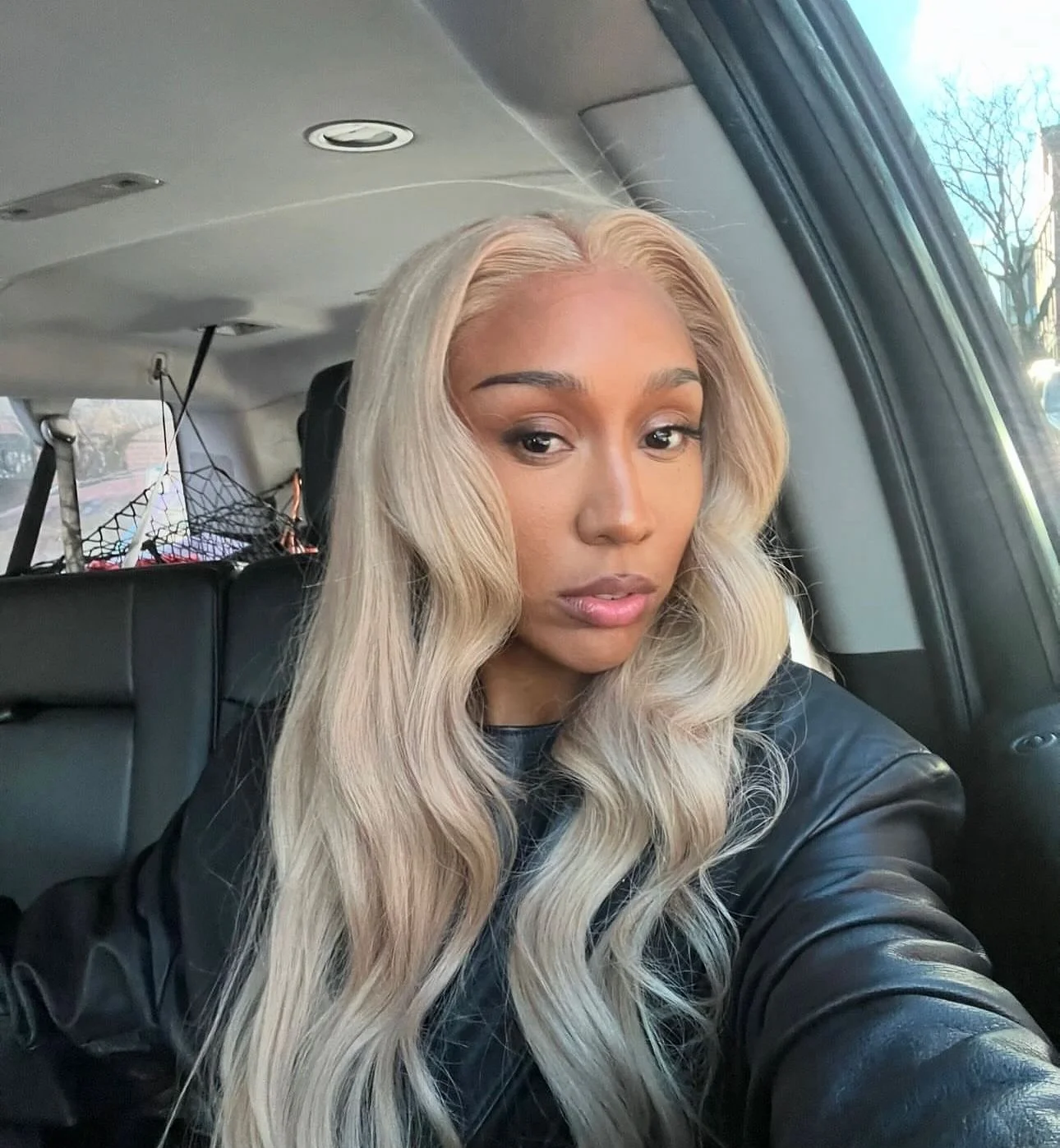 A woman with long, wavy blonde hair taking a selfie inside a vehicle, with a black leather jacket.