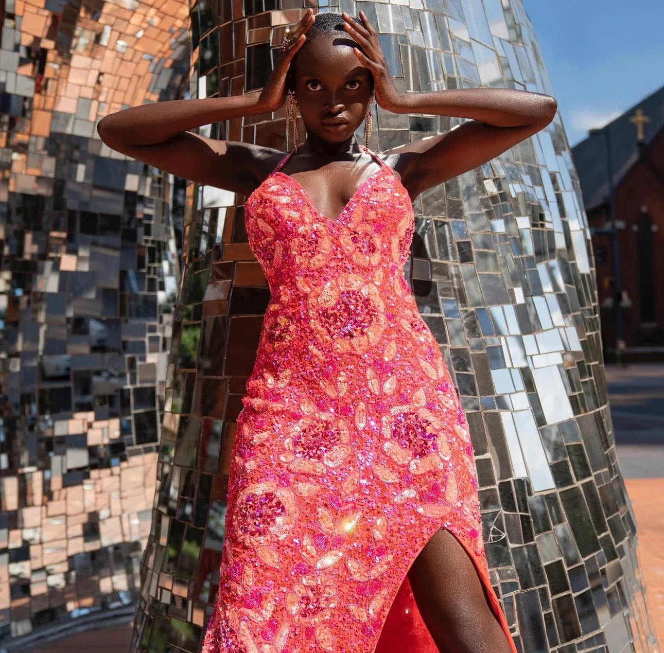 A woman in a vibrant pink sequin dress with a high slit, standing outdoors against a mirrored sculpture and city buildings.