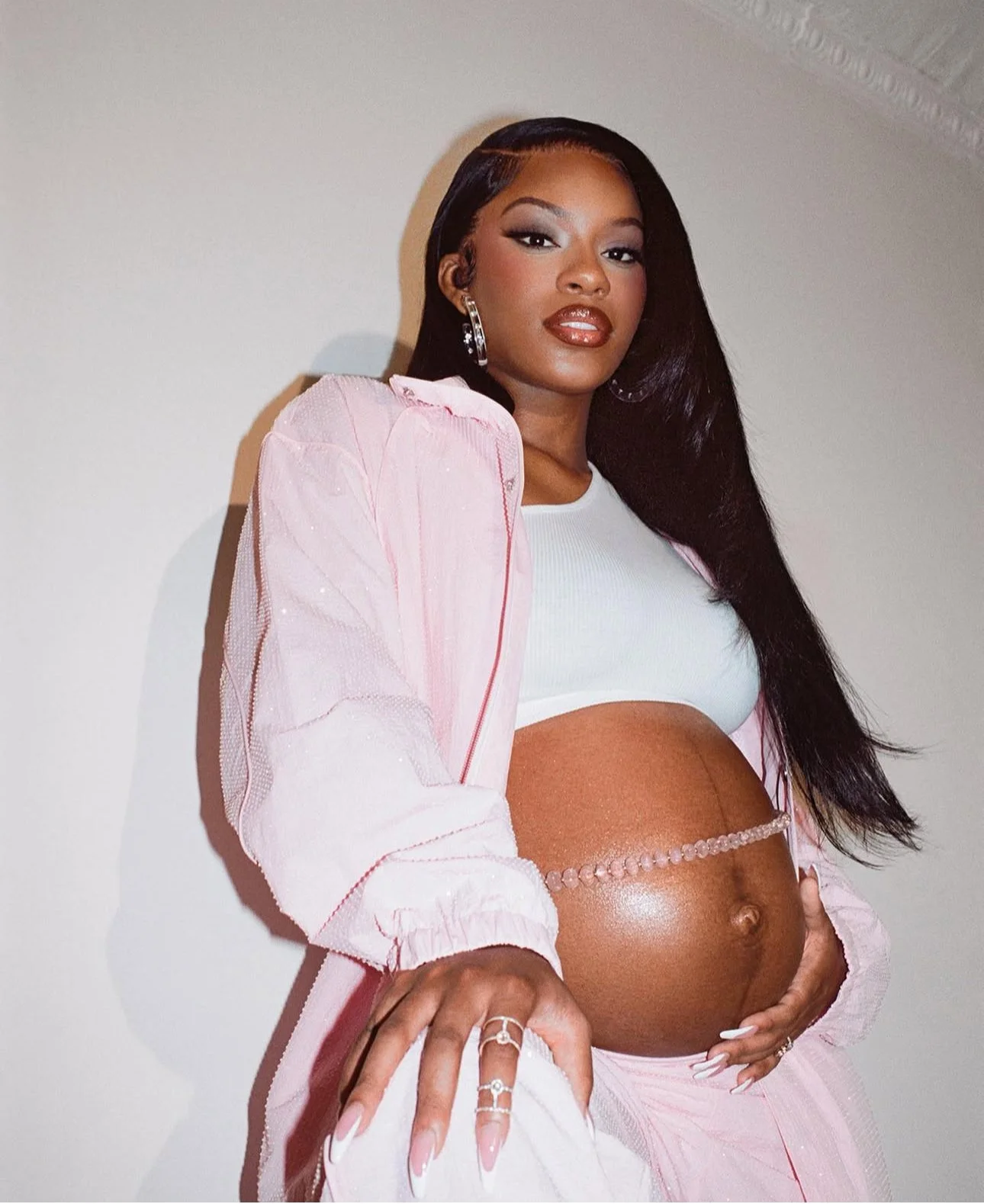 A pregnant woman with long dark hair, wearing a white crop top, pink jacket, and pink pants, standing against a plain white wall.