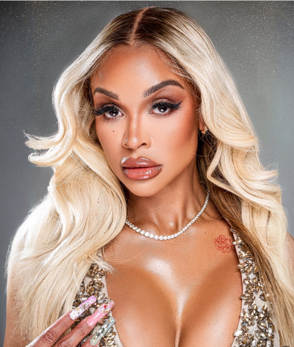 A woman with long blonde hair, makeup, and a pearl necklace, holding her decorated hand to her chest, wearing a sparkling top.