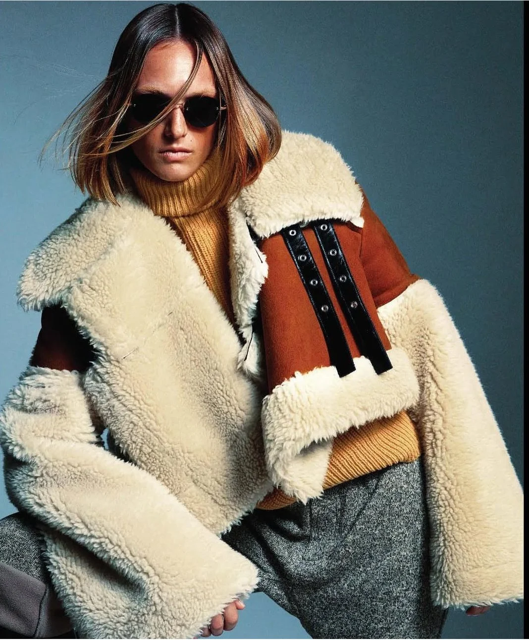 A woman wearing dark sunglasses, a yellow turtleneck sweater, and a shearling-lined jacket with brown, black, and cream colors, posing against a gray background.