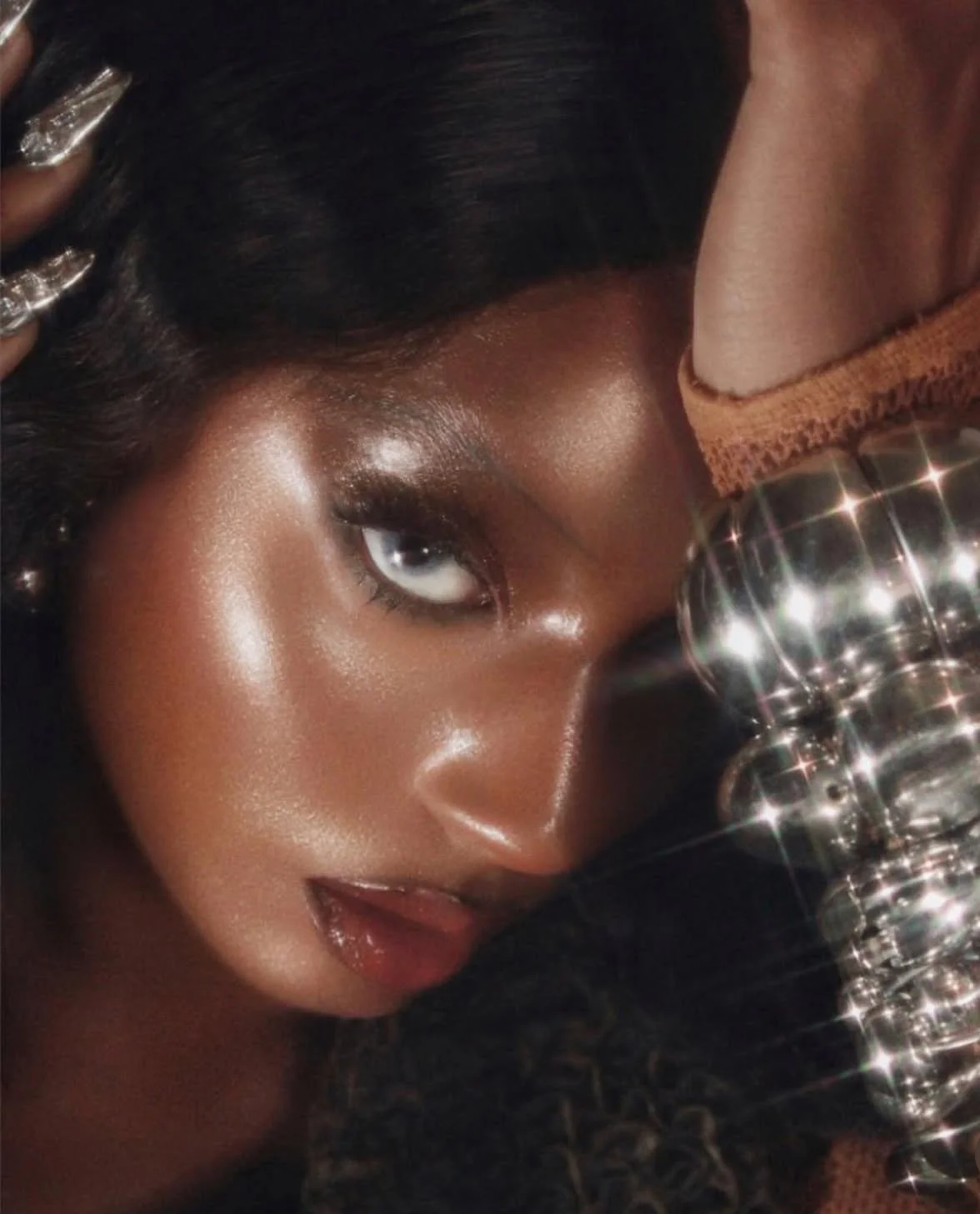 A close-up photo of a woman with shiny makeup, dark hair, and sparkly jewelry, lying down with her hand in her hair, next to a cluster of shiny glasses.