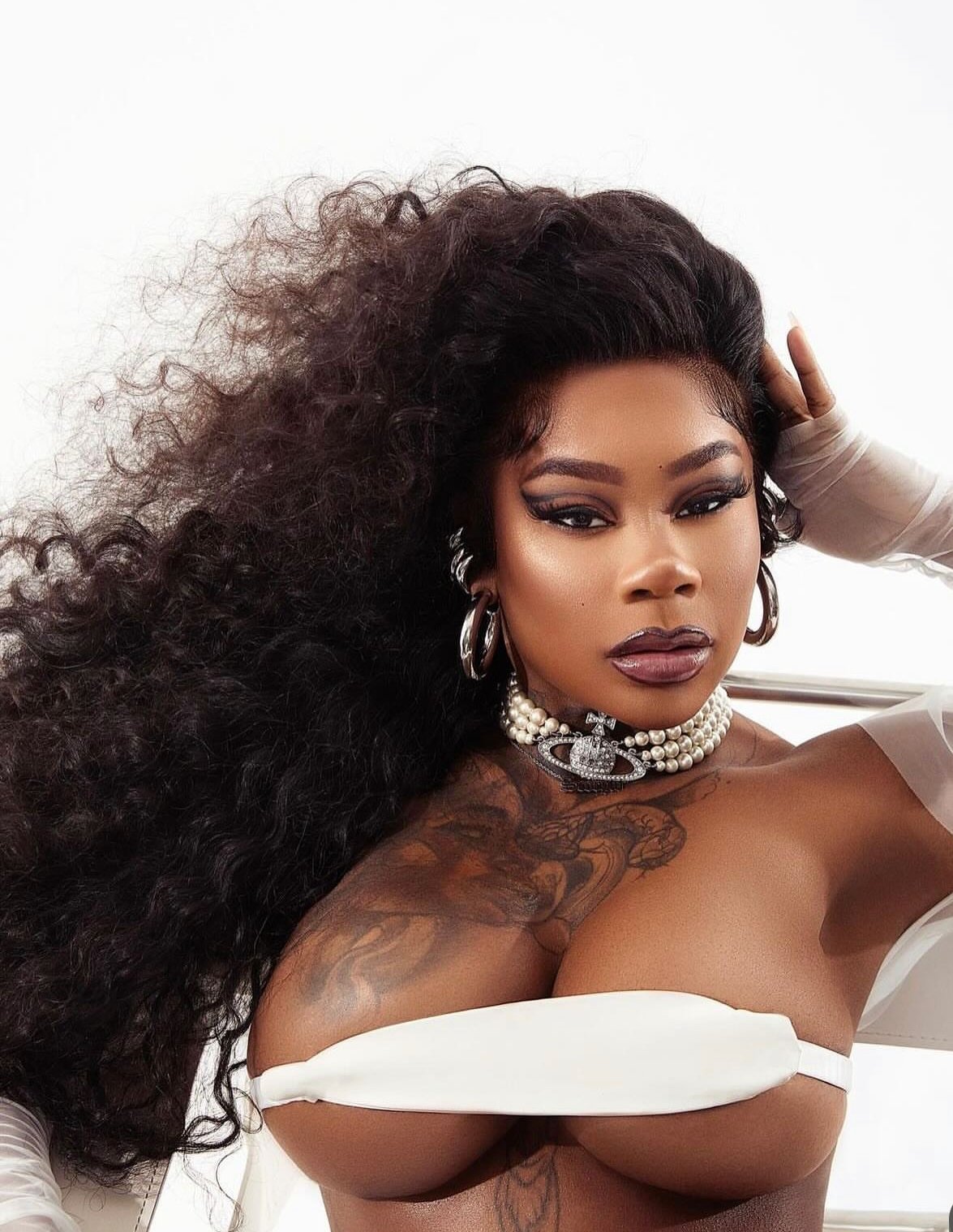 A woman with long, curly black hair, wearing large hoop earrings, a pearl necklace with a crown pendant, and dark makeup, posing against a white background.