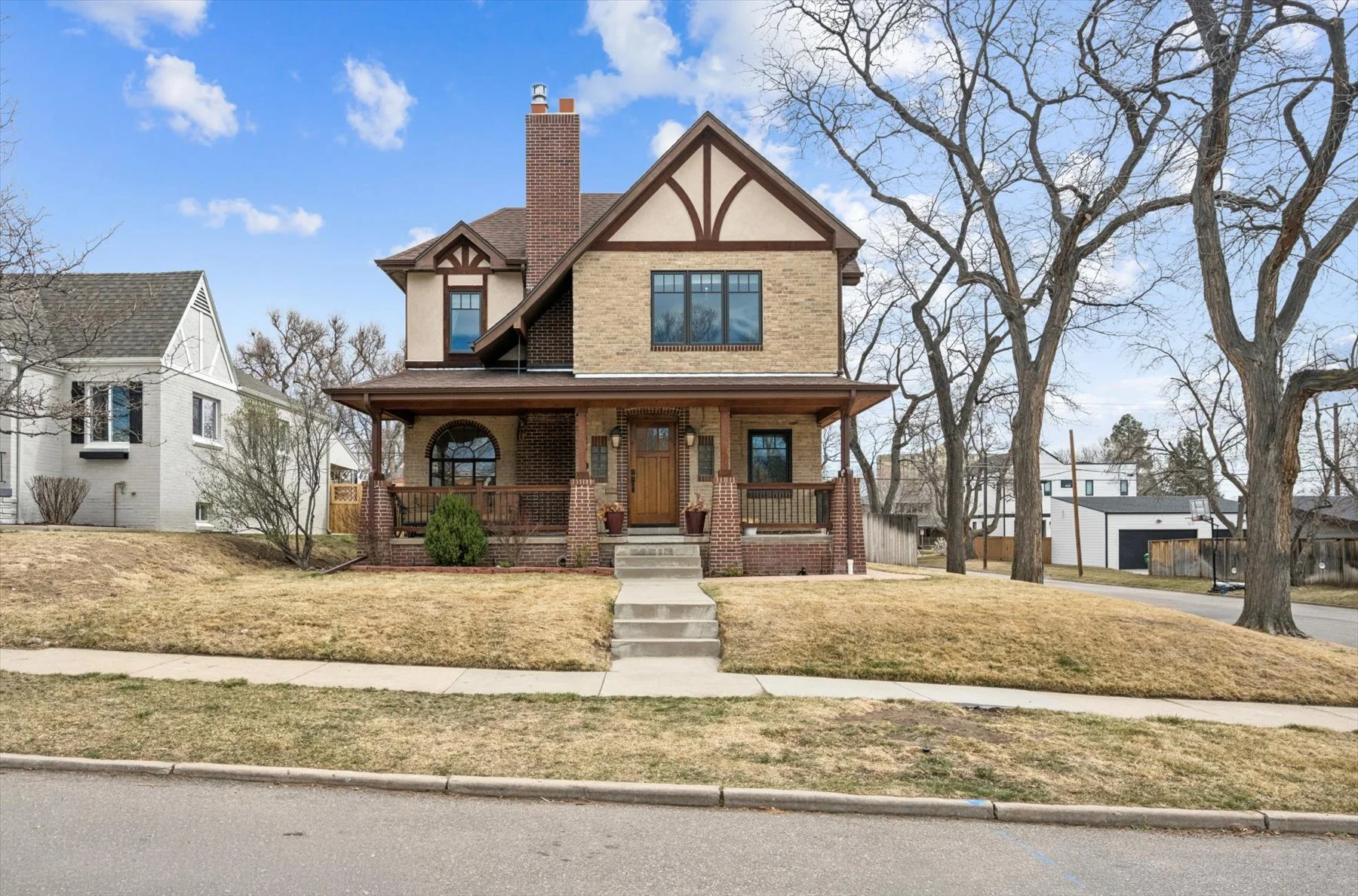A two-story house with a front porch, brick and beige siding, and a gabled roof. There are stairs leading to the porch, and trees with no leaves are visible around the house.