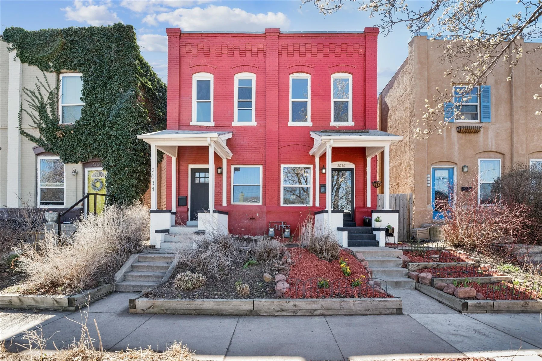Two-story red brick row house with white trim, black front door, small front porch, and front yard with dried plants and rocks, situated between two other houses with similar style, in a neighborhood with bare trees and a sidewalk.