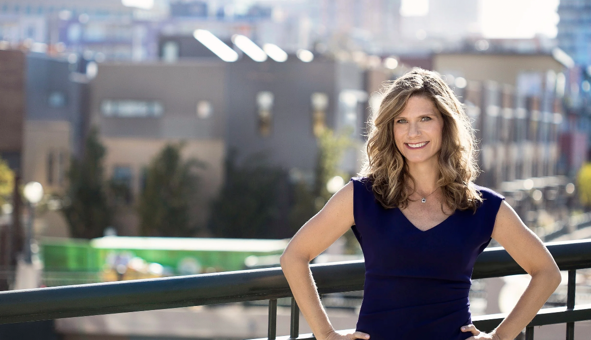 A smiling woman with wavy, shoulder-length blonde hair standing outdoors on a balcony with a cityscape in the background.