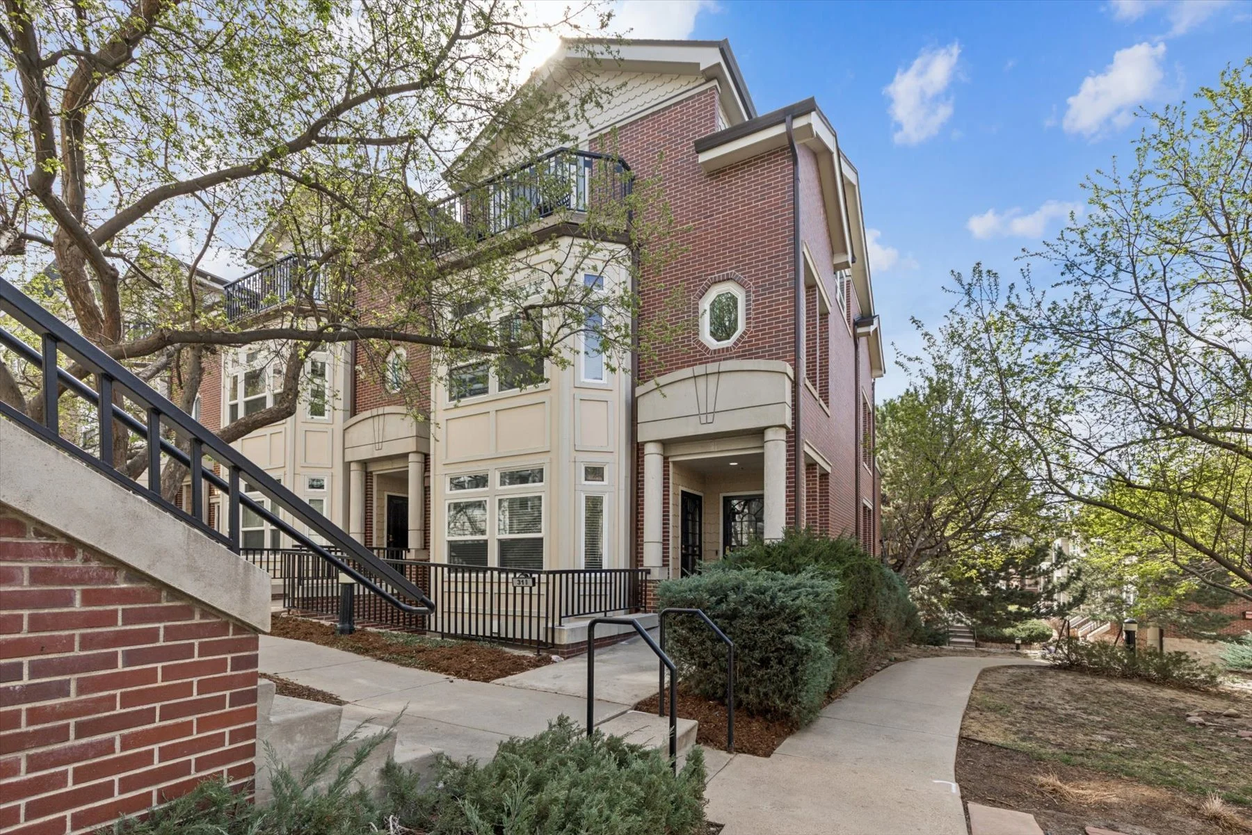 Multi-story brick residential building with large windows, a small balcony, and a front entrance, surrounded by trees and a landscaped walkway.