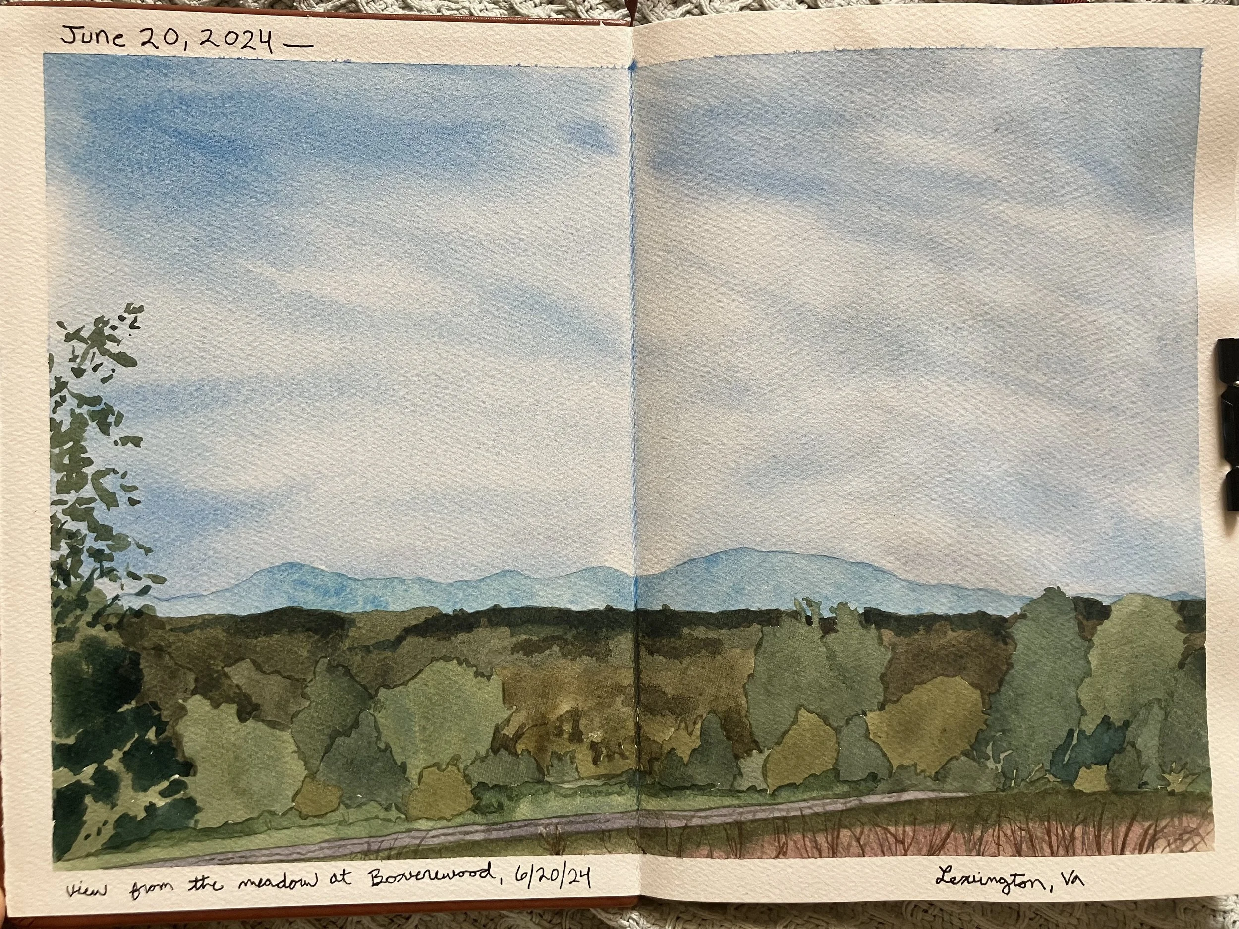 A watercolor painting of a landscape with a cloudy sky, distant mountains, and a foreground with green bushes and a red-brown field. The date June 20, 2024, is written at the top left, and notes at the bottom indicate the view is from the meadow at Boverwood in Lexington, VA, on June 20, 2024.