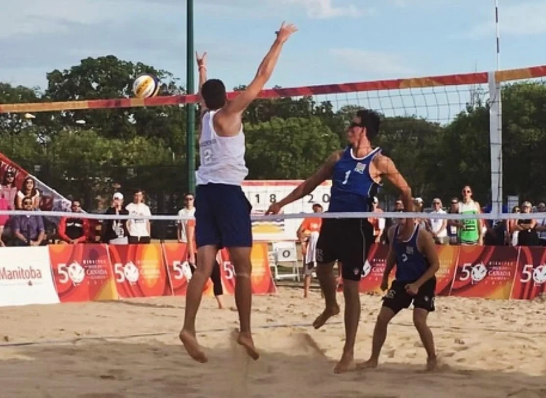 Two male beach volleyball players in blue and white uniforms are jumping to hit the volleyball at the net during a match, with spectators watching in the background.
