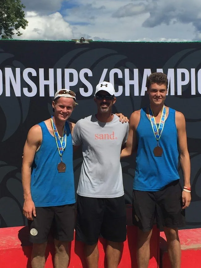 Two young male athletes wearing blue tank tops and medals around their necks stand with an adult male in a white t-shirt with 'sand.' written on it, in front of a backdrop with partially visible text 'CHAMPIONSHIPS.' The photo was taken outdoors during daytime.