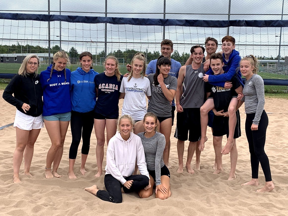 Group of young athletes posing on a sand volleyball court outside, with some wearing athletic clothing and others in casual clothes, smiling for the camera.