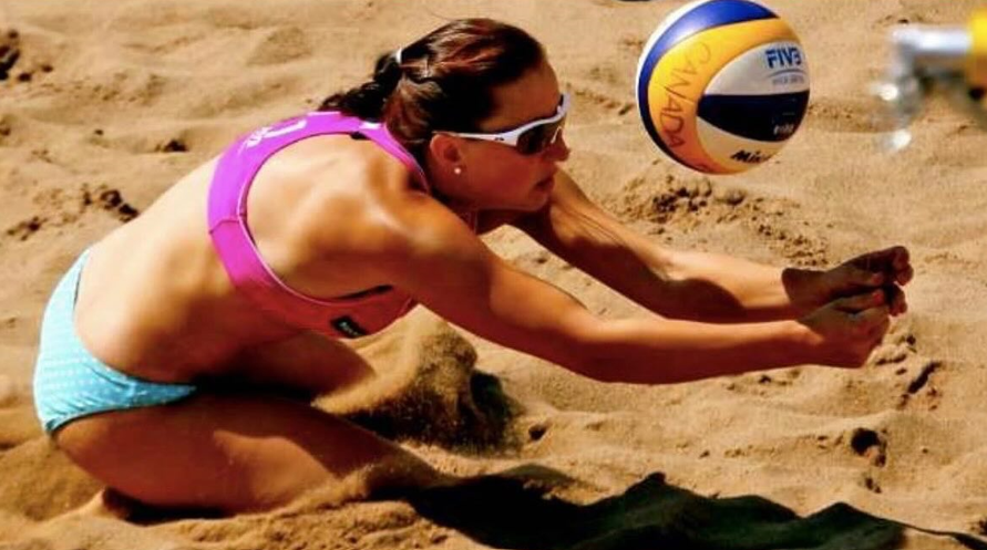 A female beach volleyball player wearing sunglasses, a pink sports top, and light blue shorts, diving into the sand to hit a volleyball.