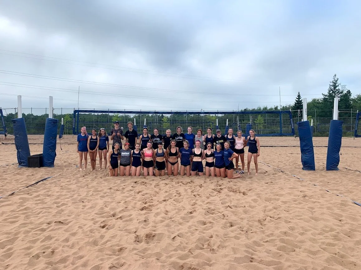 Group of young women and coaches posing after a beach volleyball game on sand court with overcast sky.