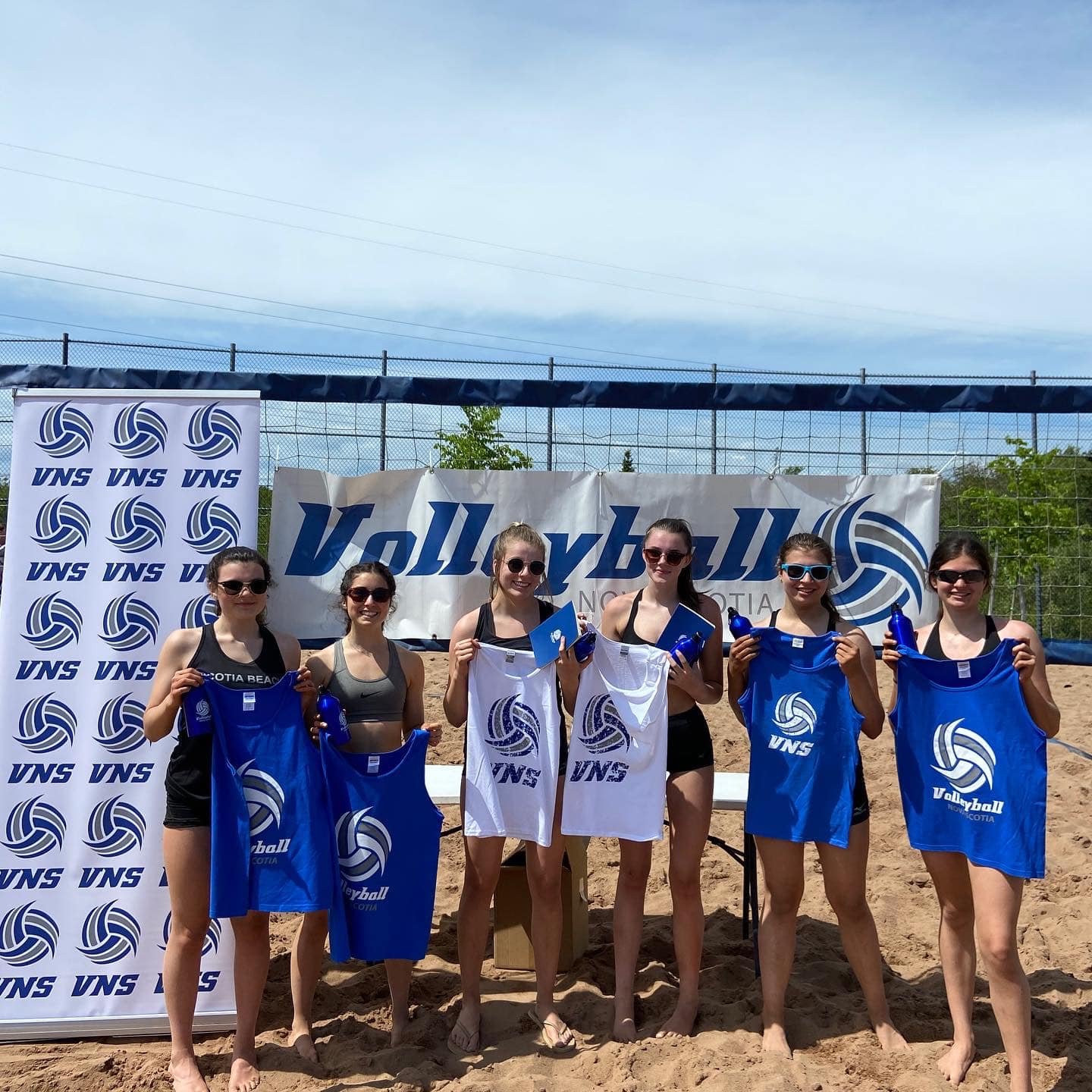 Group of six young women standing on a volleyball court, holding blue and white volleyball team shirts with the VNS logo, during a sunny day, with banners and a blue sky in the background.