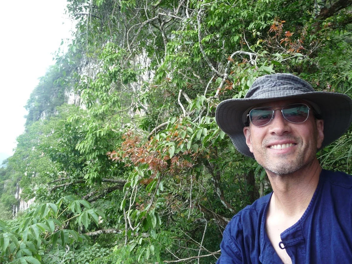 A man wearing sunglasses, a wide-brimmed hat, and a blue shirt, smiling outdoors in front of dense green foliage and trees.