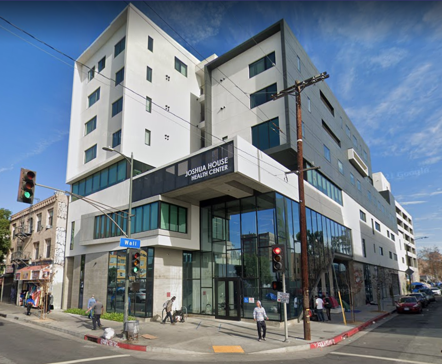 Modern multi-story building with glass storefront and sign reading 'Joshua House Health Center' on corner of a busy street.