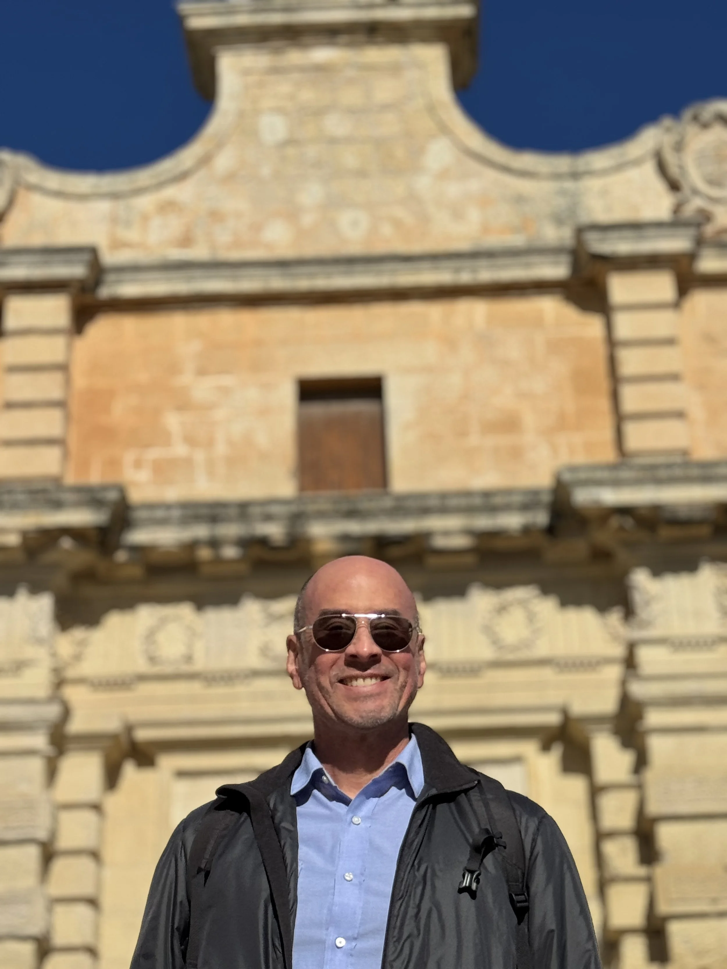 A man wearing sunglasses, a light blue button-up shirt, and a black jacket, standing and smiling in front of a historic stone building with architectural details.
