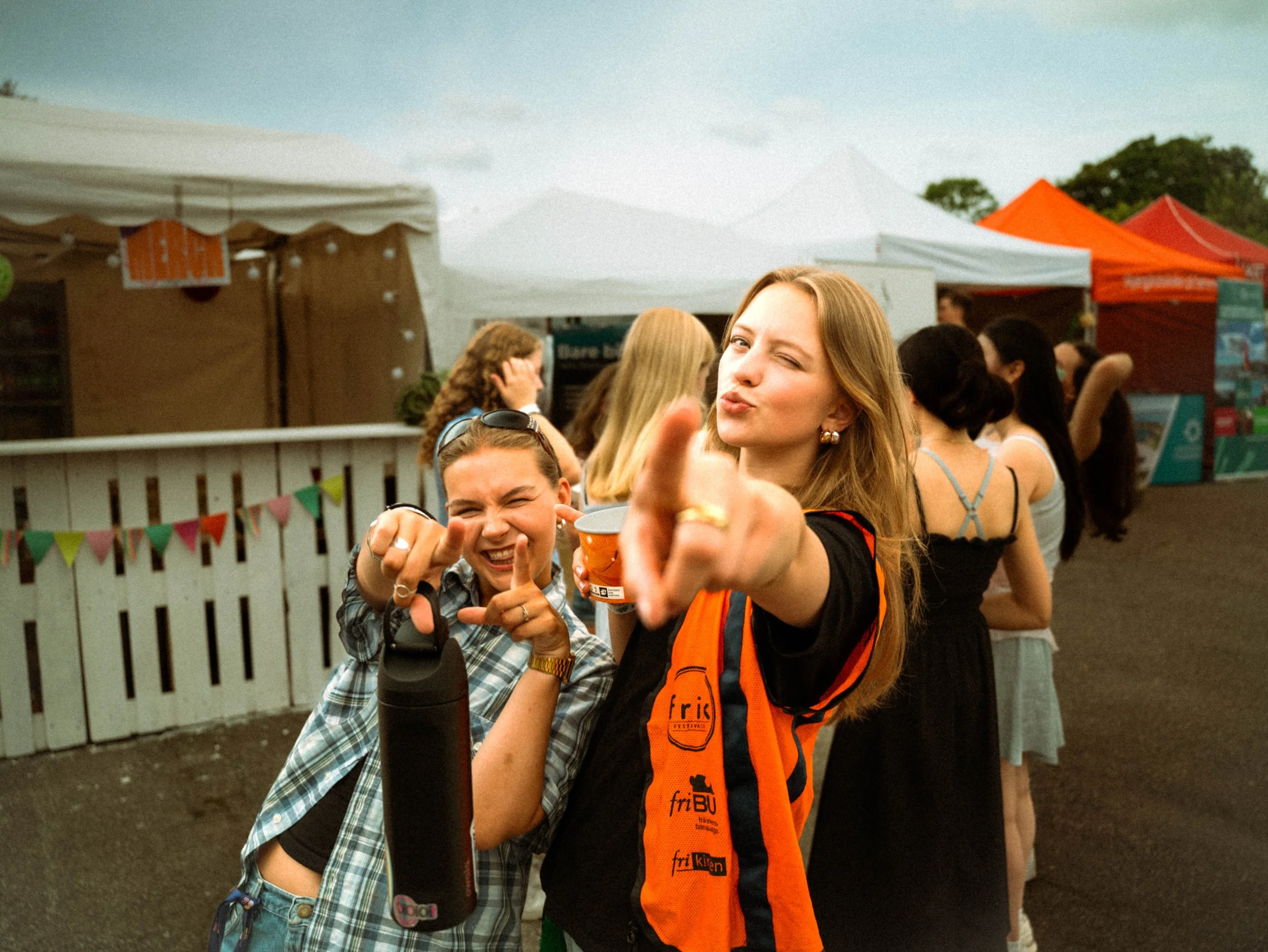 To unge kvinner som smiler og peker mot kamera under en utendørs festival eller marked, med flere andre mennesker og fargerike telt i bakgrunnen.