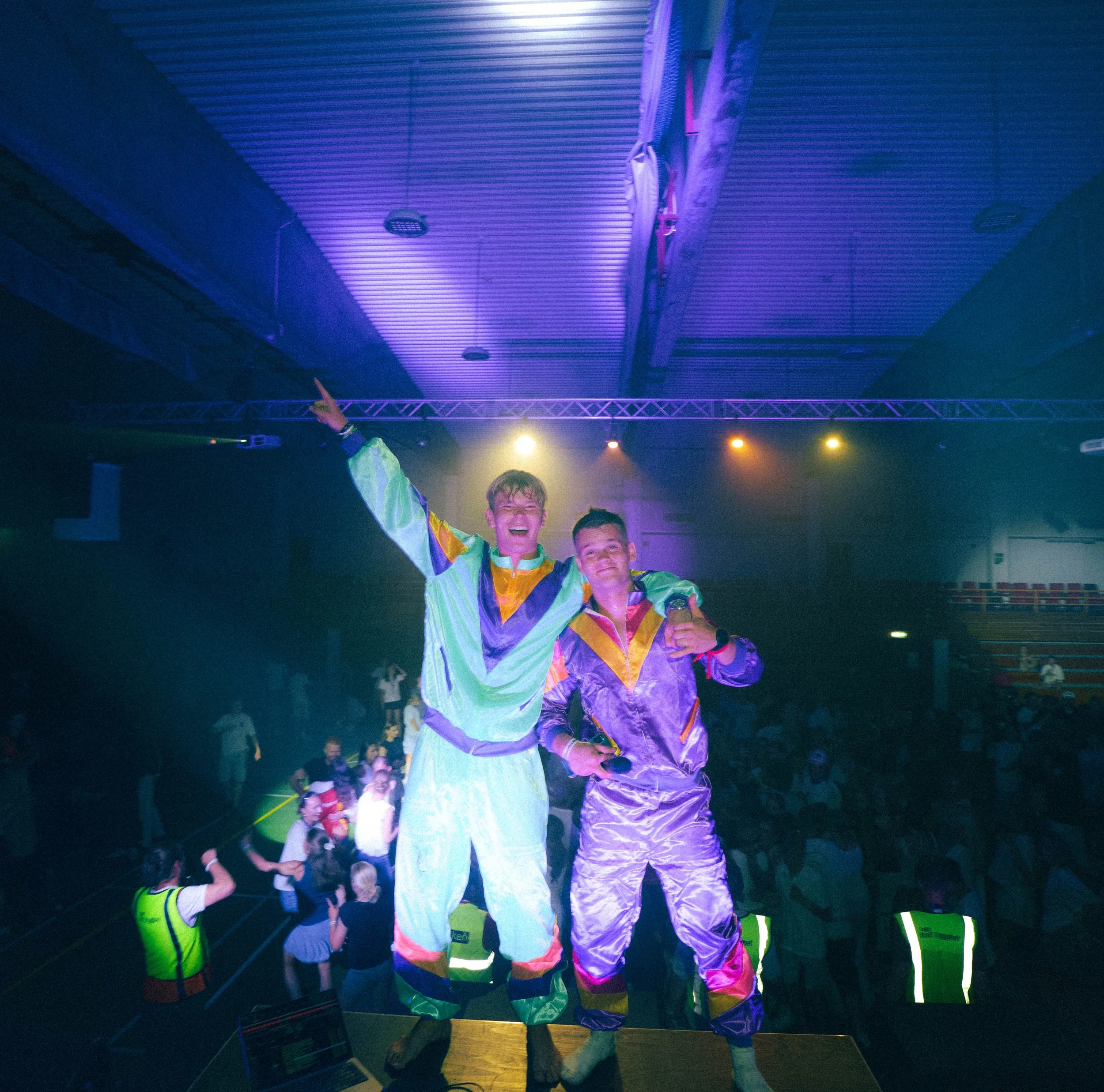To personer i fargerike dansedrakter på en scene med folk og sikkerhetsvakter i bakgrunnen, under lyssetting i en innendørshall.