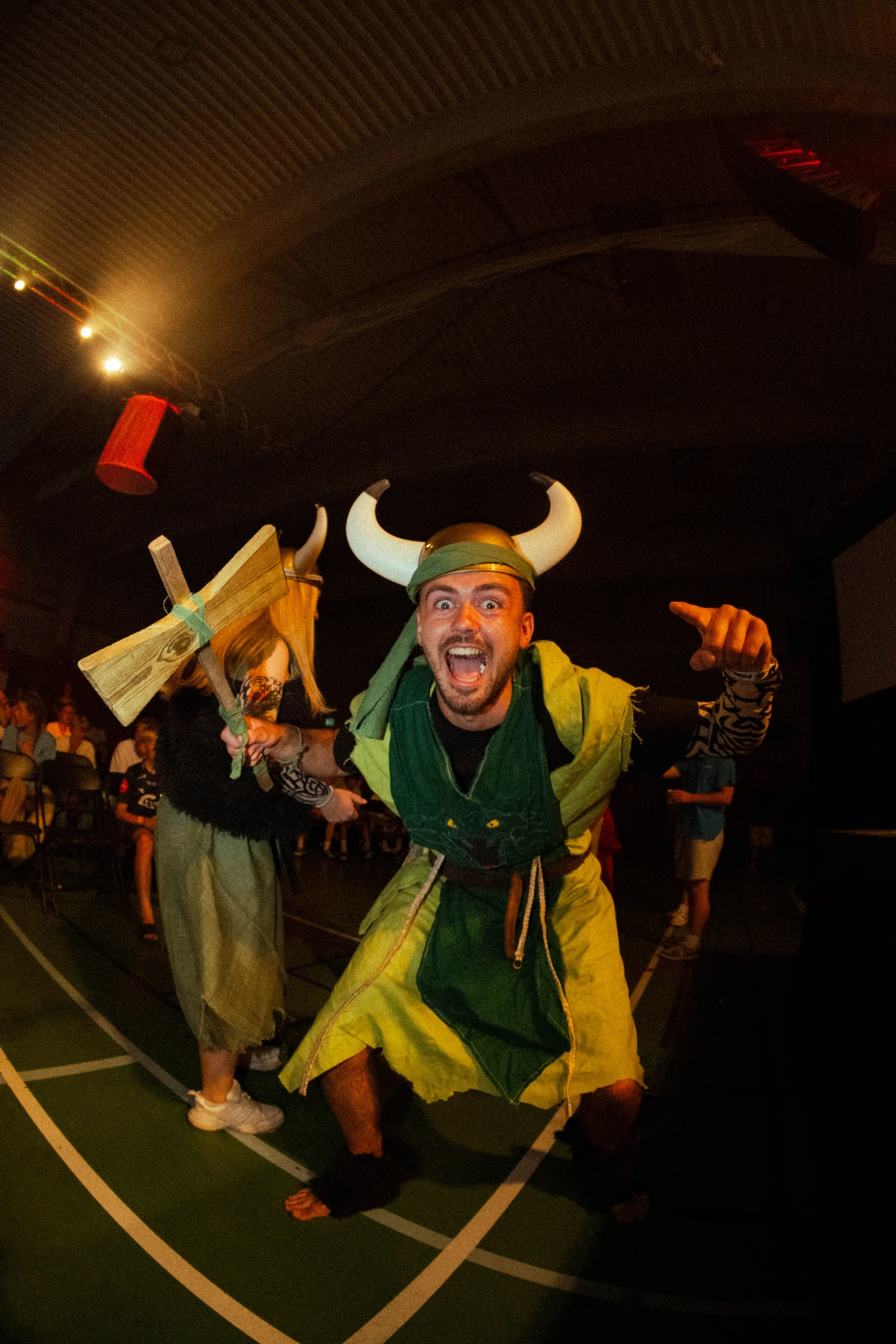 Person kledd som viking med hjelm og skjegg smilende og peker foran, mens annen person i bakgrunnen holder et spyd og har på seg en drakt.