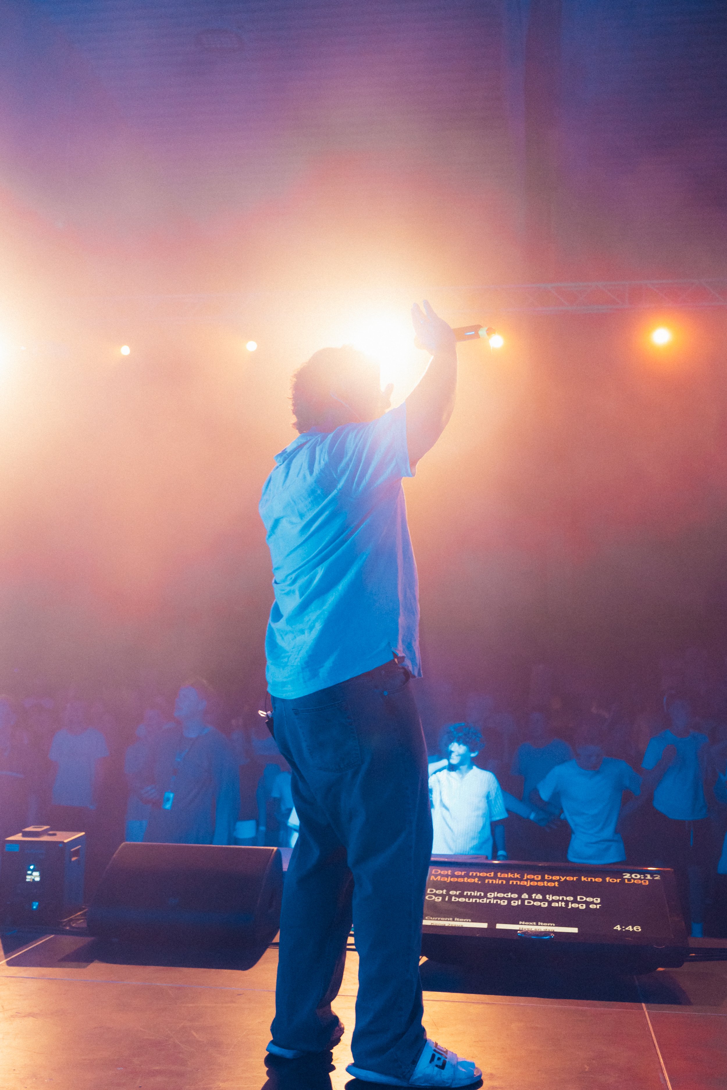 En person står på en scene og holder en mikrofon opp i luften, omgitt av fargede scenelys, med en gruppe mennesker i bakgrunnen.