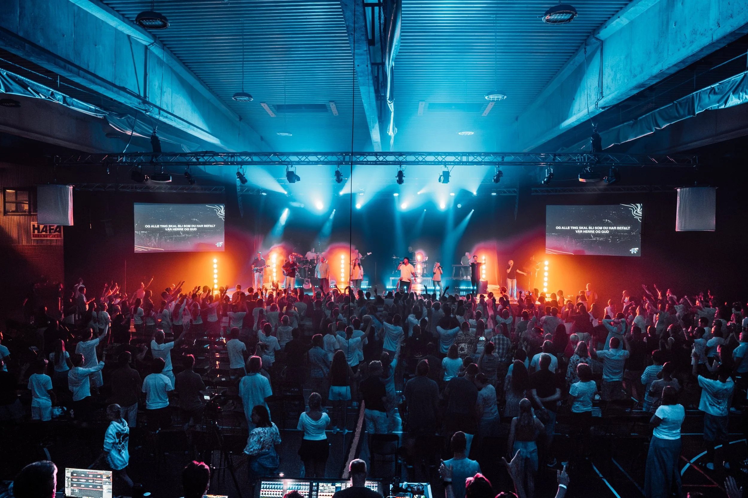 En konsert i en stor innendørs arena med en stor mengde folk som peker og danser, med scenelys i blå og oransje. Artister opptrer på scenen, og det er to store skjermer som viser tekst på norsk.