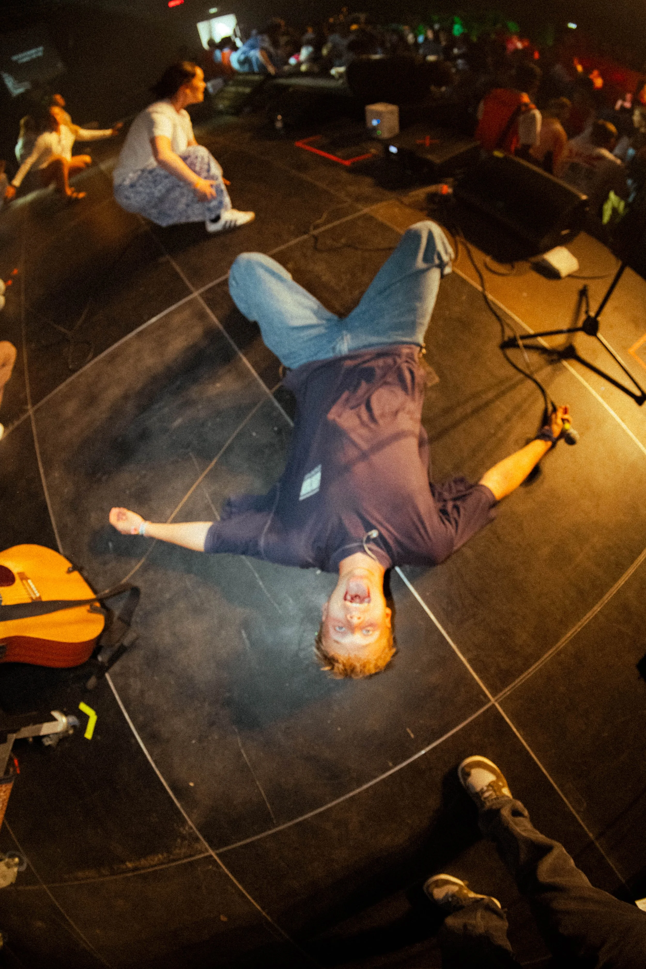 Person lying on the stage with a big smile, surrounded by musical equipment, with audience in the background.