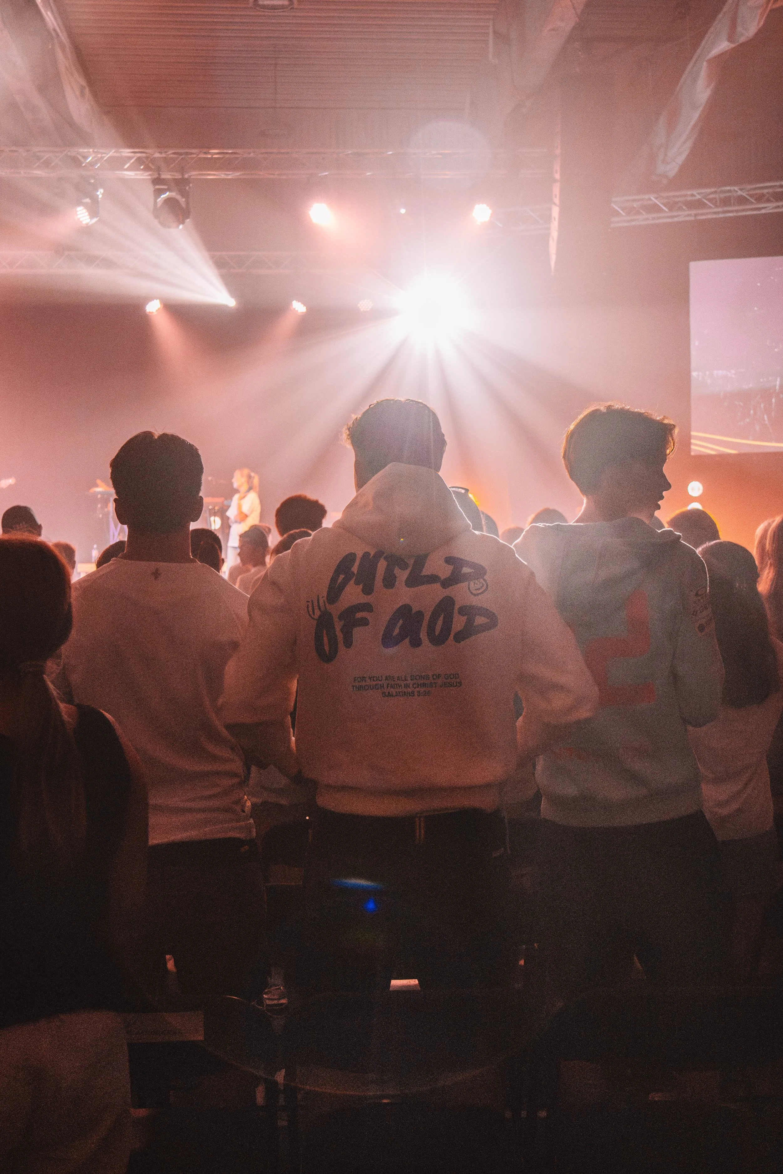 Personer ser på en scene under en konsert eller event, med lysene og en stor skjerm i bakgrunnen, i et innendørs lokale.