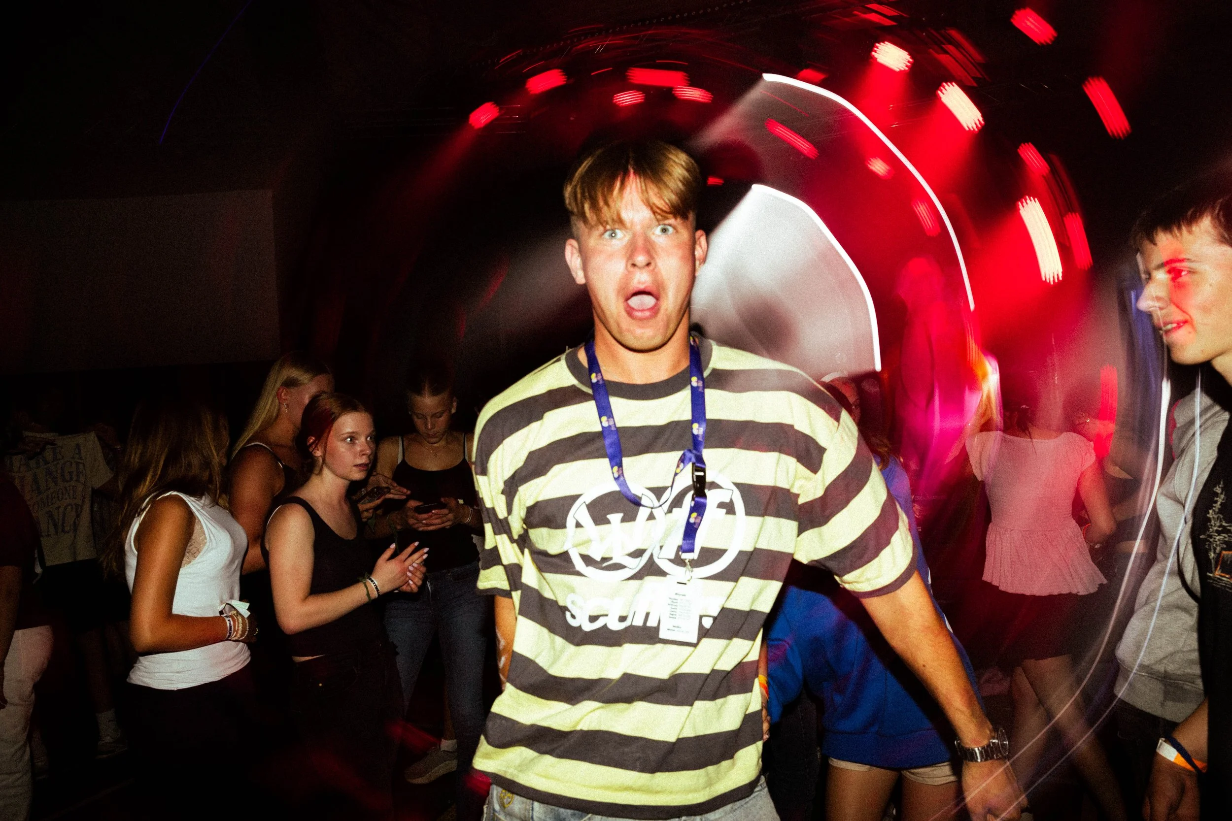 En mann i en svart-hvit stripete t-skjorte som står på dansegulvet i en klubb, omgitt av flere personer, med rødt og hvitt lys som skaper bevegelsesuskarphet.