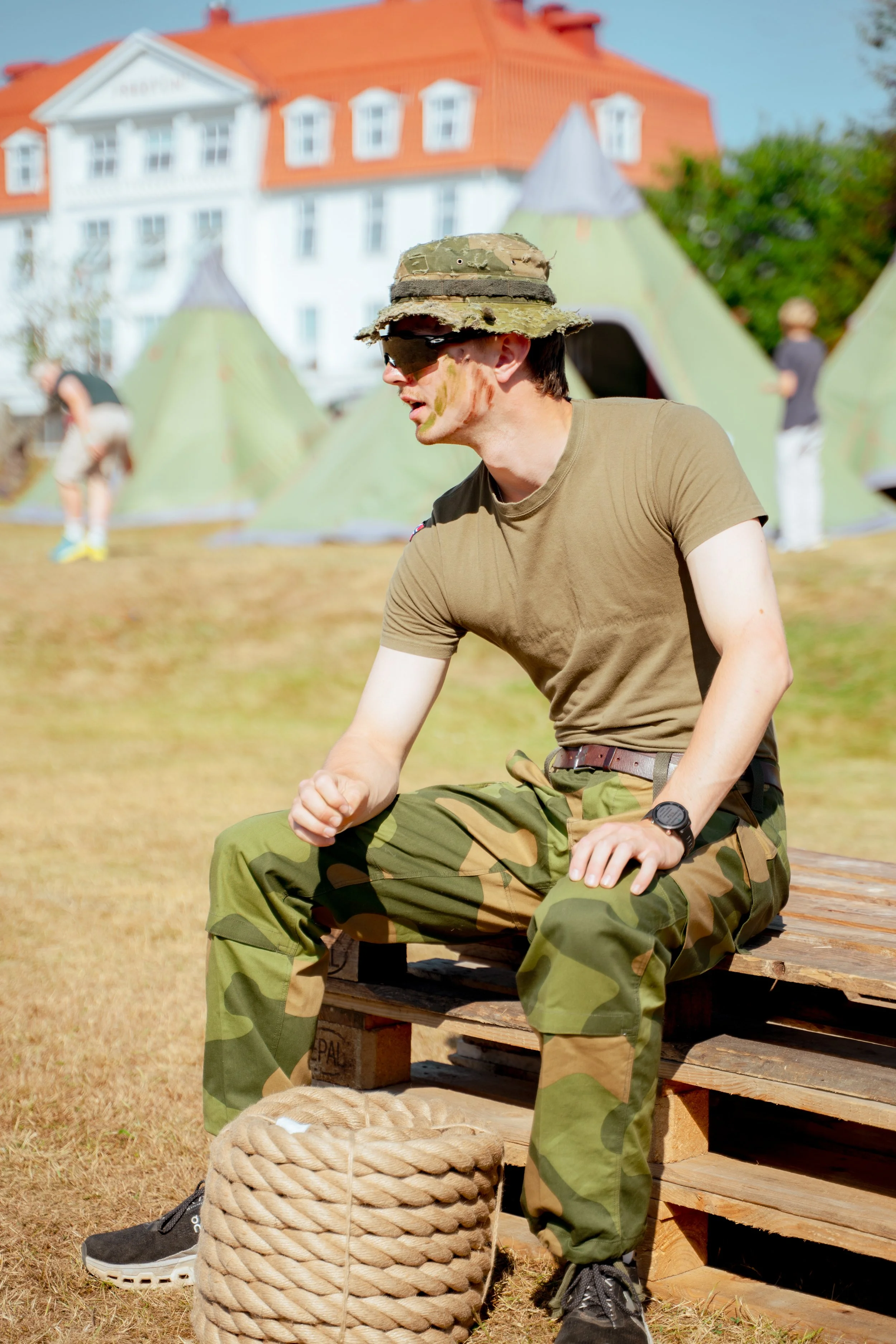 En person i soldatuniform med kylling- eller kamuflasjekledd hatt, sitter på en treskinne i et utendørs område med telt i bakgrunnen.