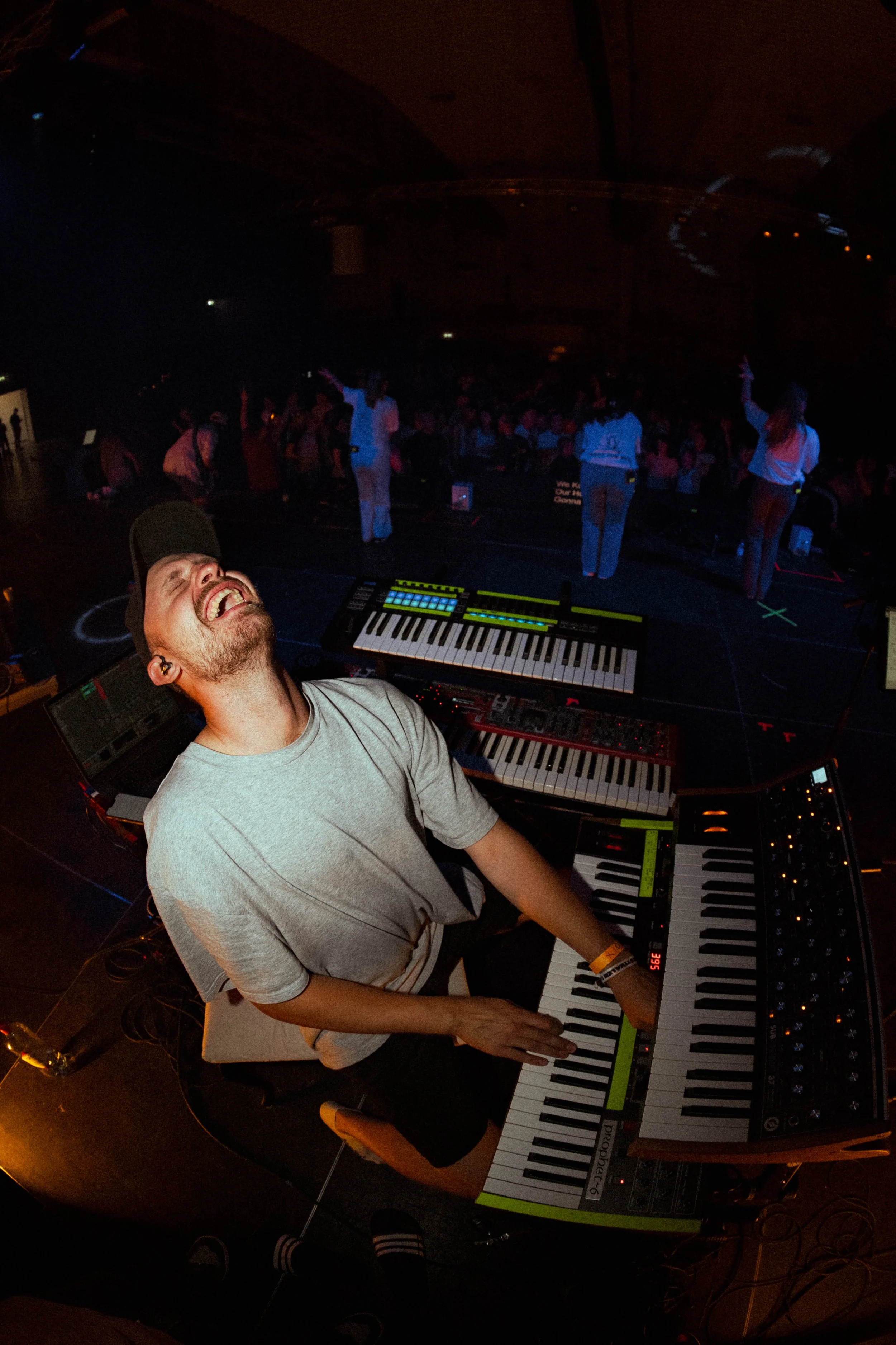 En mann med caps ler og spiller på synthesizere under en konsert. Publikum danser i bakgrunnen i en mørk sal.