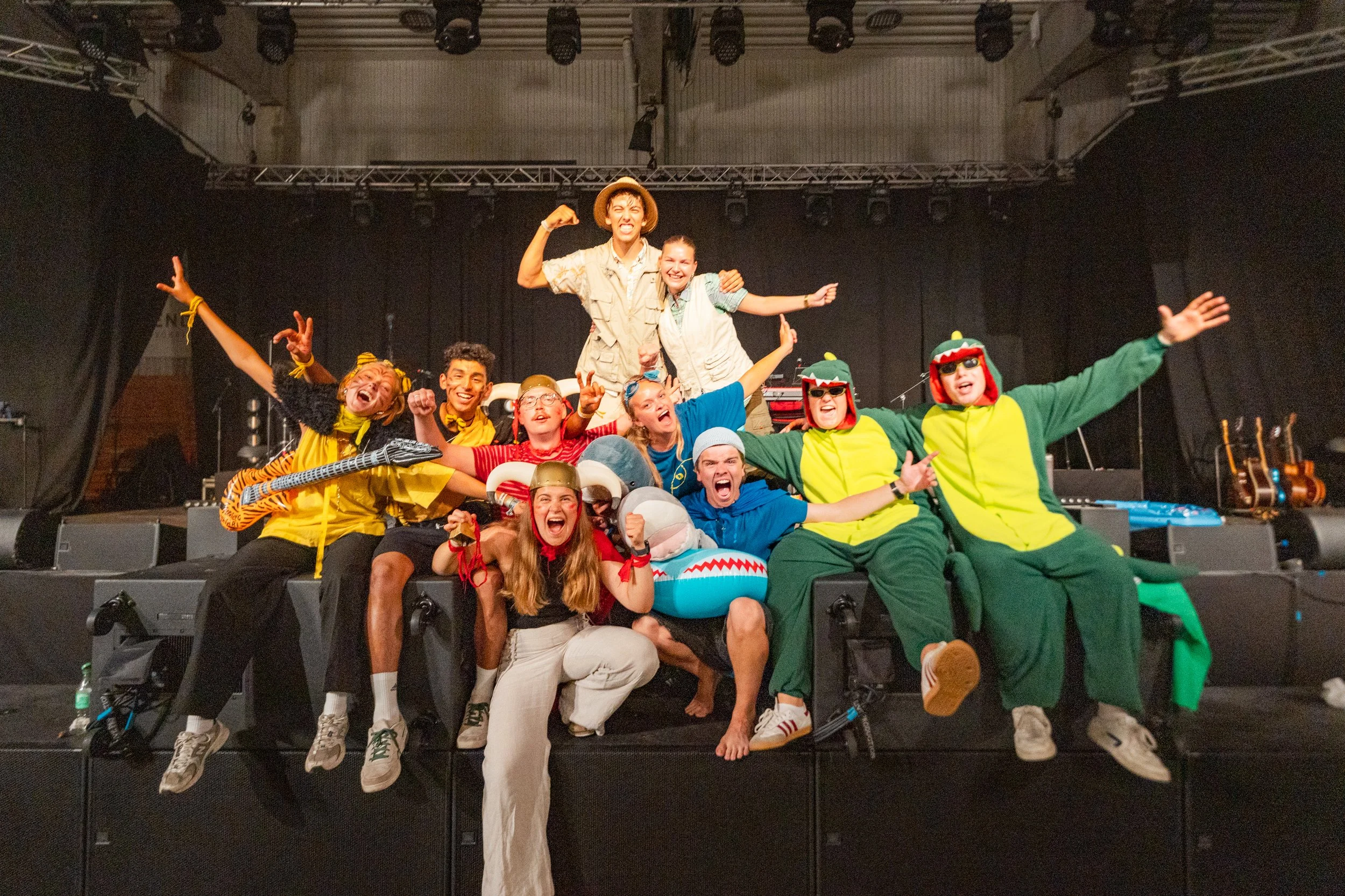En gruppe mennesker i fargerike kostymer, smilende og leende foran en svart scenetekstil, med musikkinstrumenter i bakgrunnen.