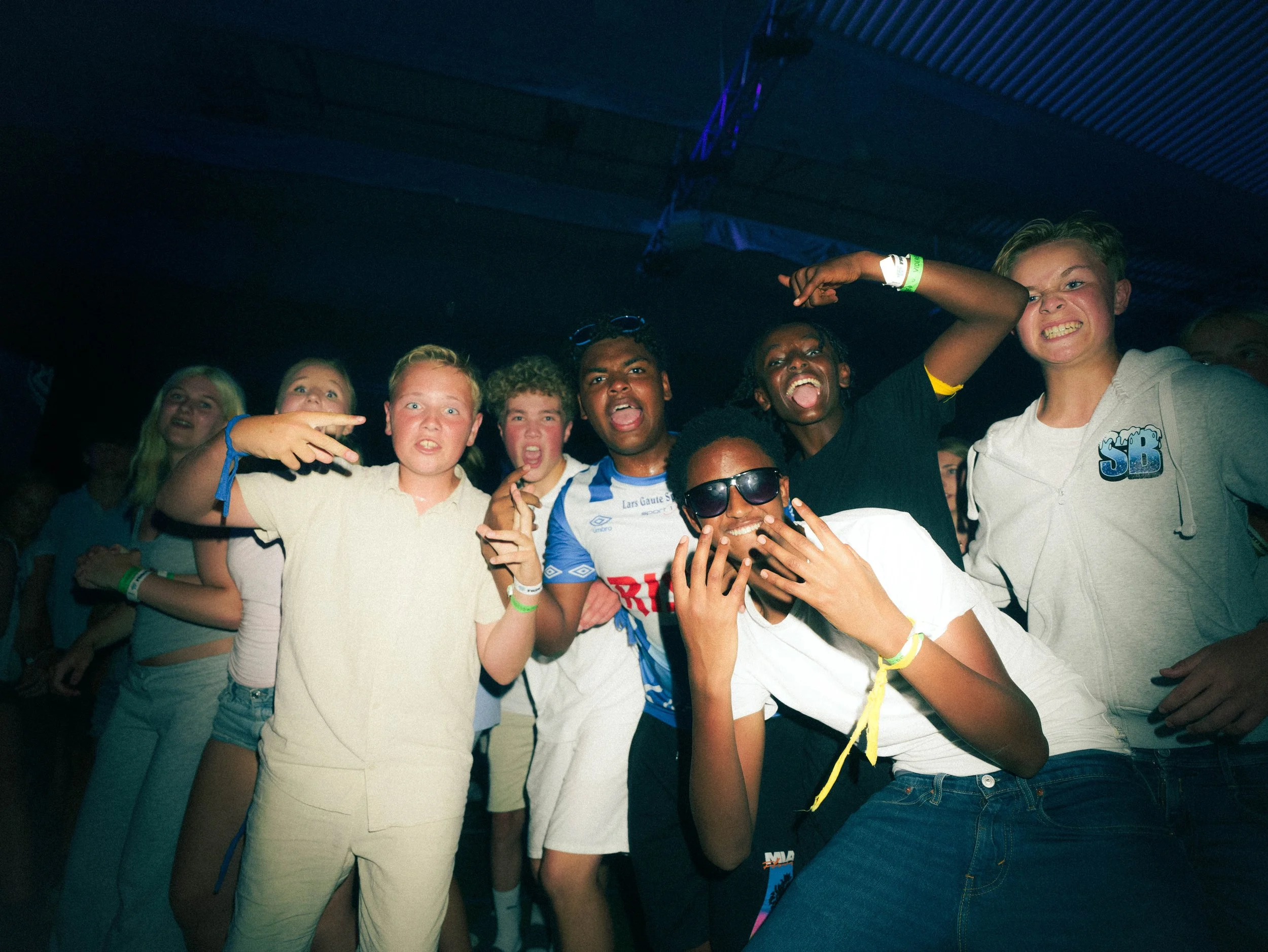 En gruppe unge mennesker som danser og har det moro på en konsert eller festival, med flere av dem som gjør seirende eller energiske håndbevegelser.