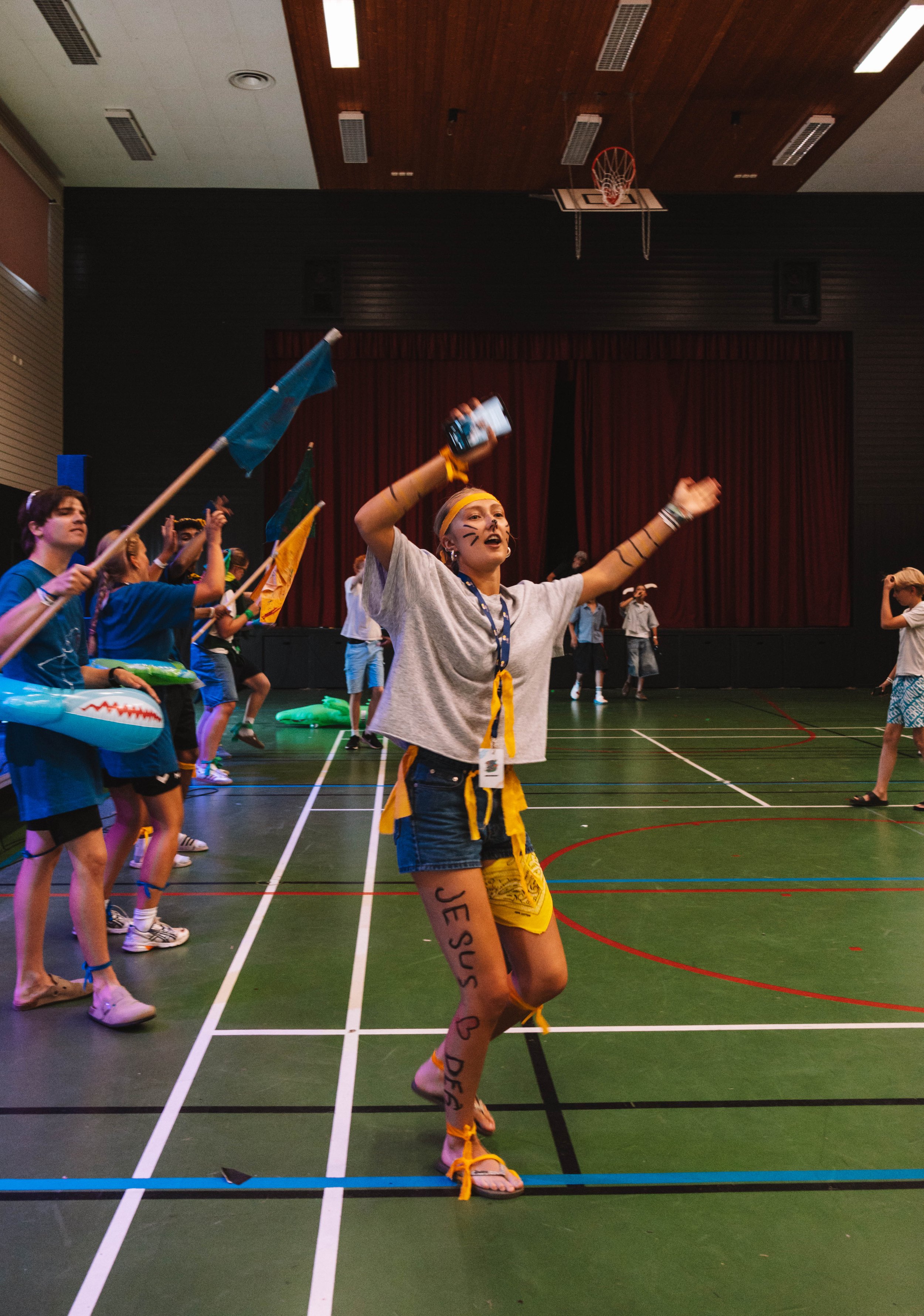 Ungdommer i en gymsal som feirer, noen med flagg og vannflasker. En jente foran har kroppstatovering med teksten «JESUS ♥ DGF» og er kledd i t-skjorte, shorts og sandaler.