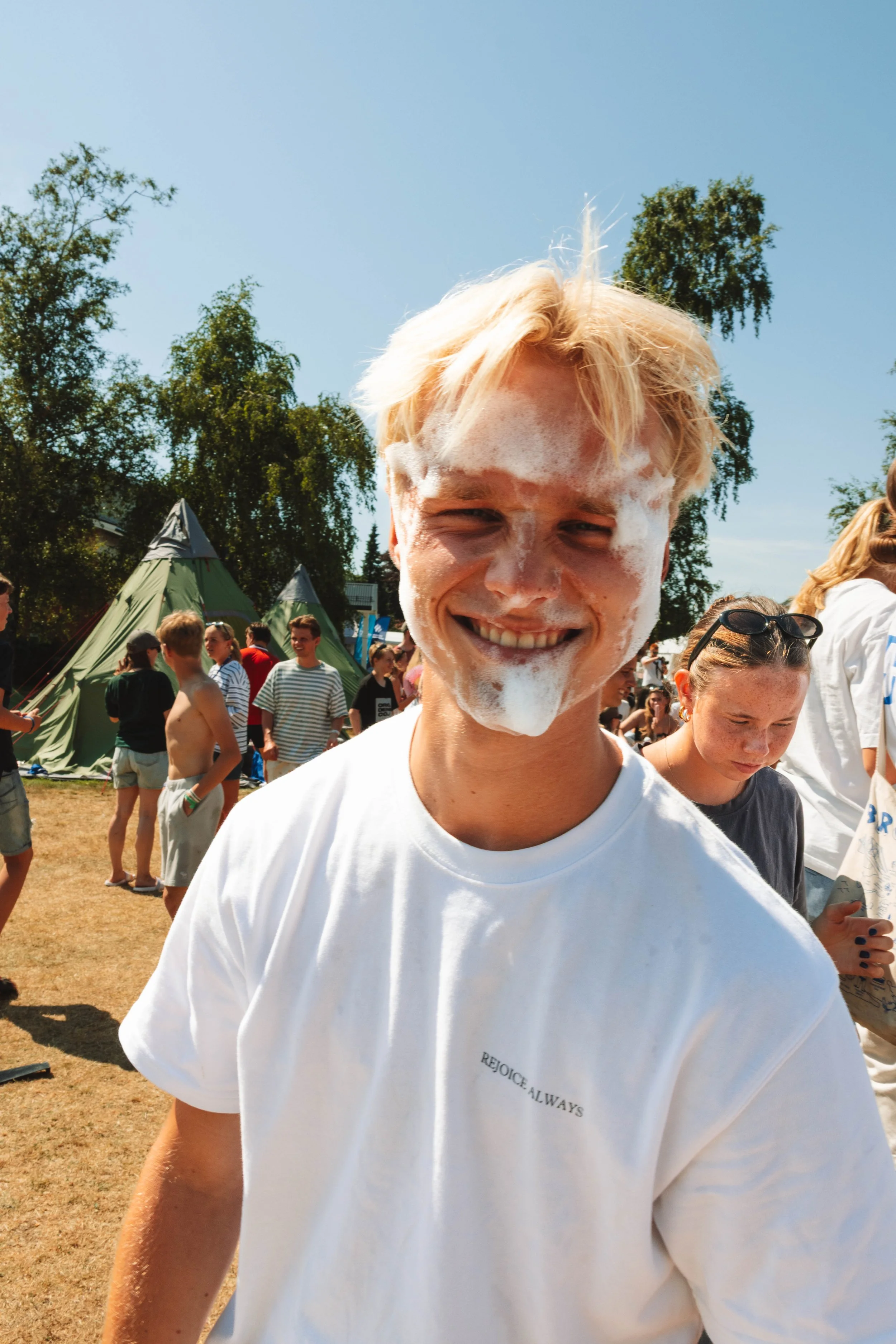 En smilende ung mann med blond hår, dekket av såpe, på en utendørs festival eller leircamp med flere mennesker og telt i bakgrunnen.